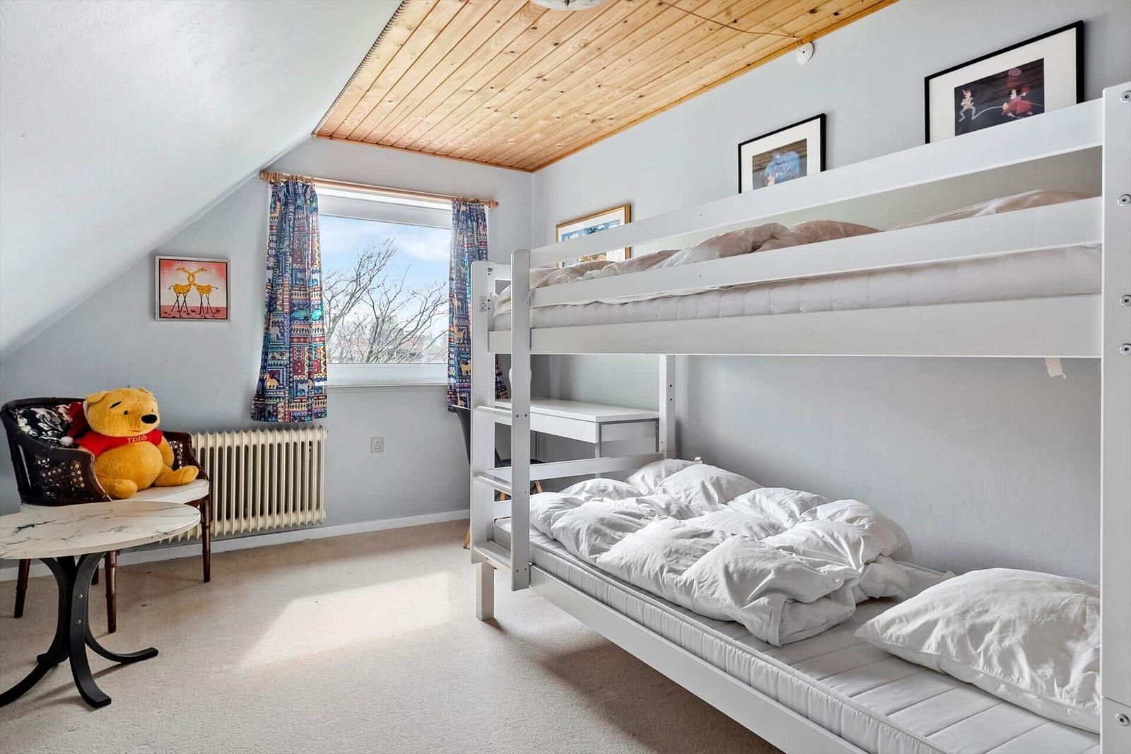 Zimmer mit Doppelstockbett, Fenster, Vorhängen und Stuhl mit Plüschfigur.