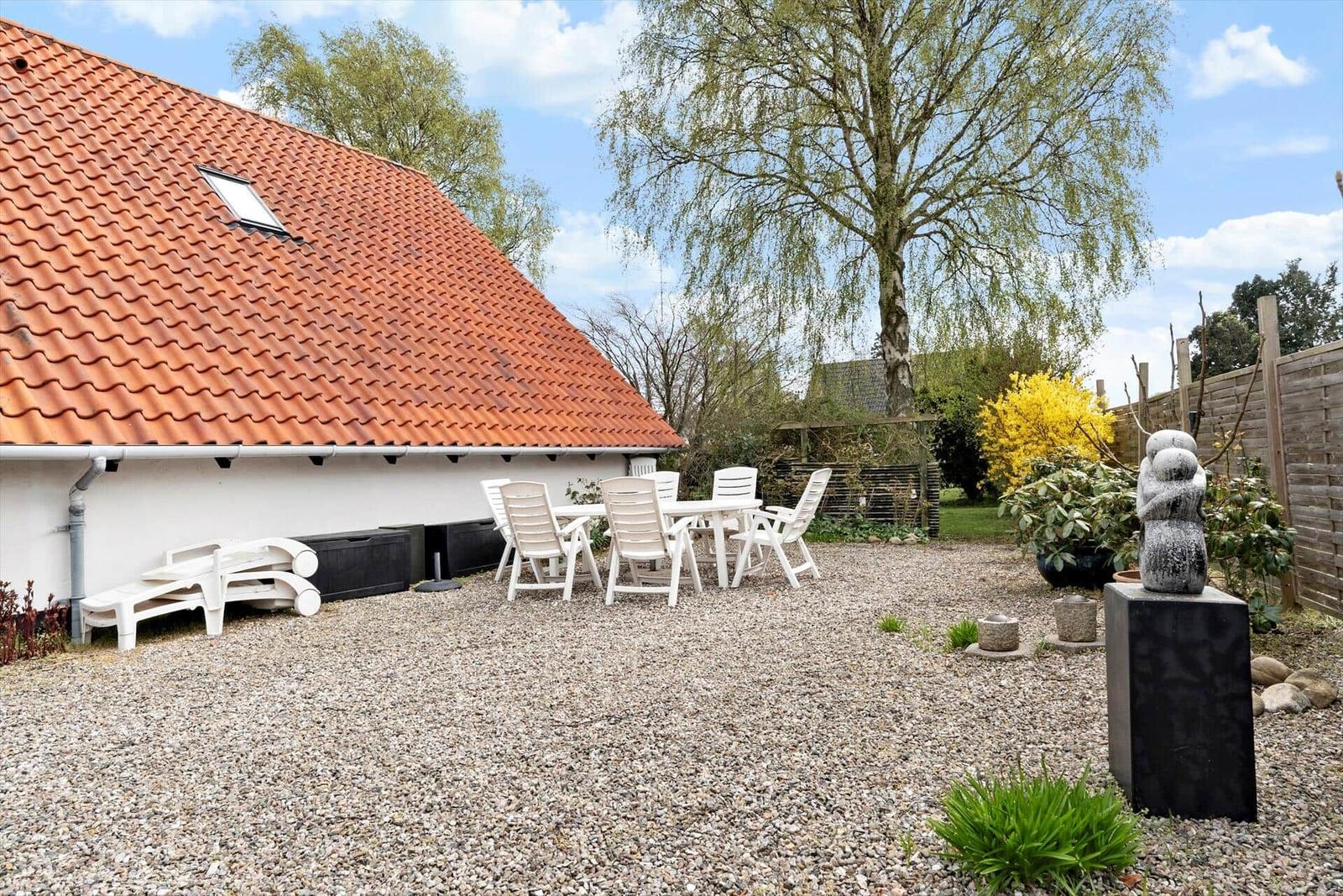Hinterhof mit Tisch, Stühlen, Steinfigur und Grasfläche unter blauem Himmel.