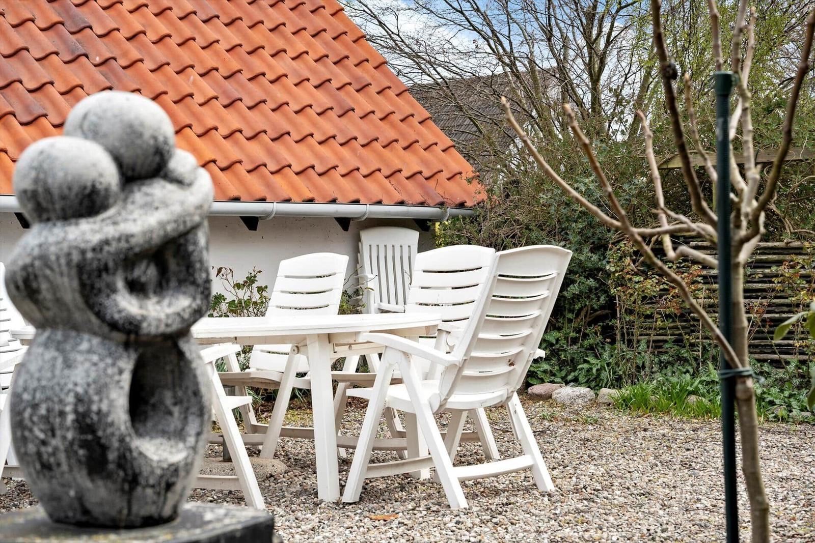 Garten mit weißem Tisch und Stühlen, Steinfigur und rotem Dach im Hintergrund.