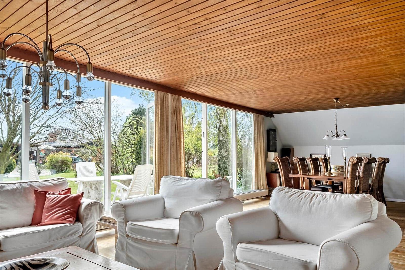 Wohn- und Essbereich mit weißen Sesseln, Holzdecke und großen Fenstern zum Garten.