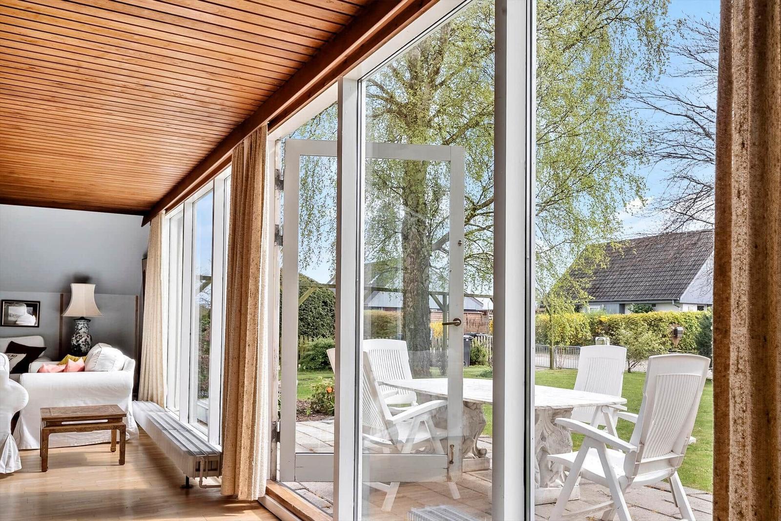 Wohnzimmer mit Holzdecke und großem Fenster zur Terrasse mit weißen Stühlen.