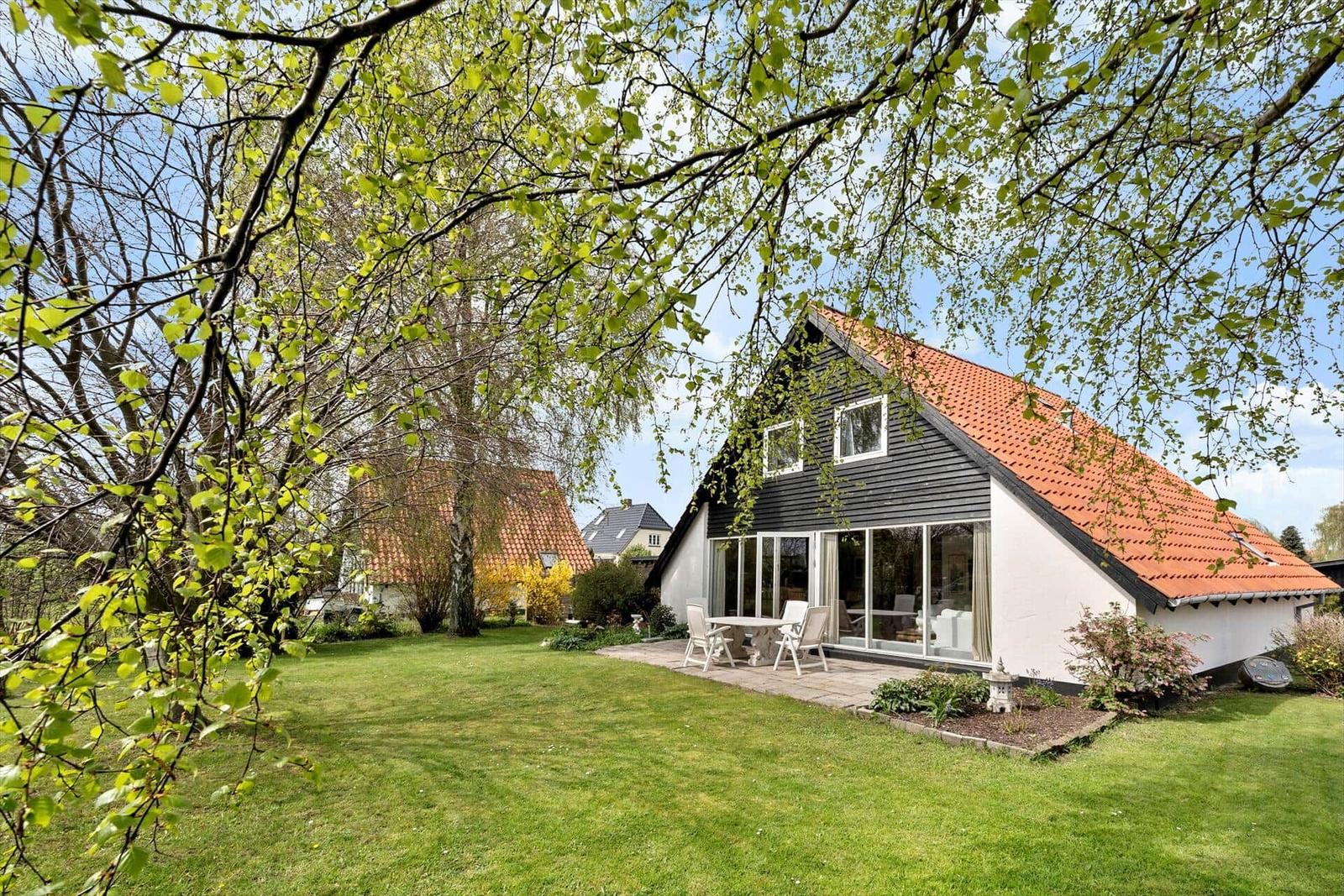Haus mit Terrasse, Garten und rotem Dach unter Bäumen.