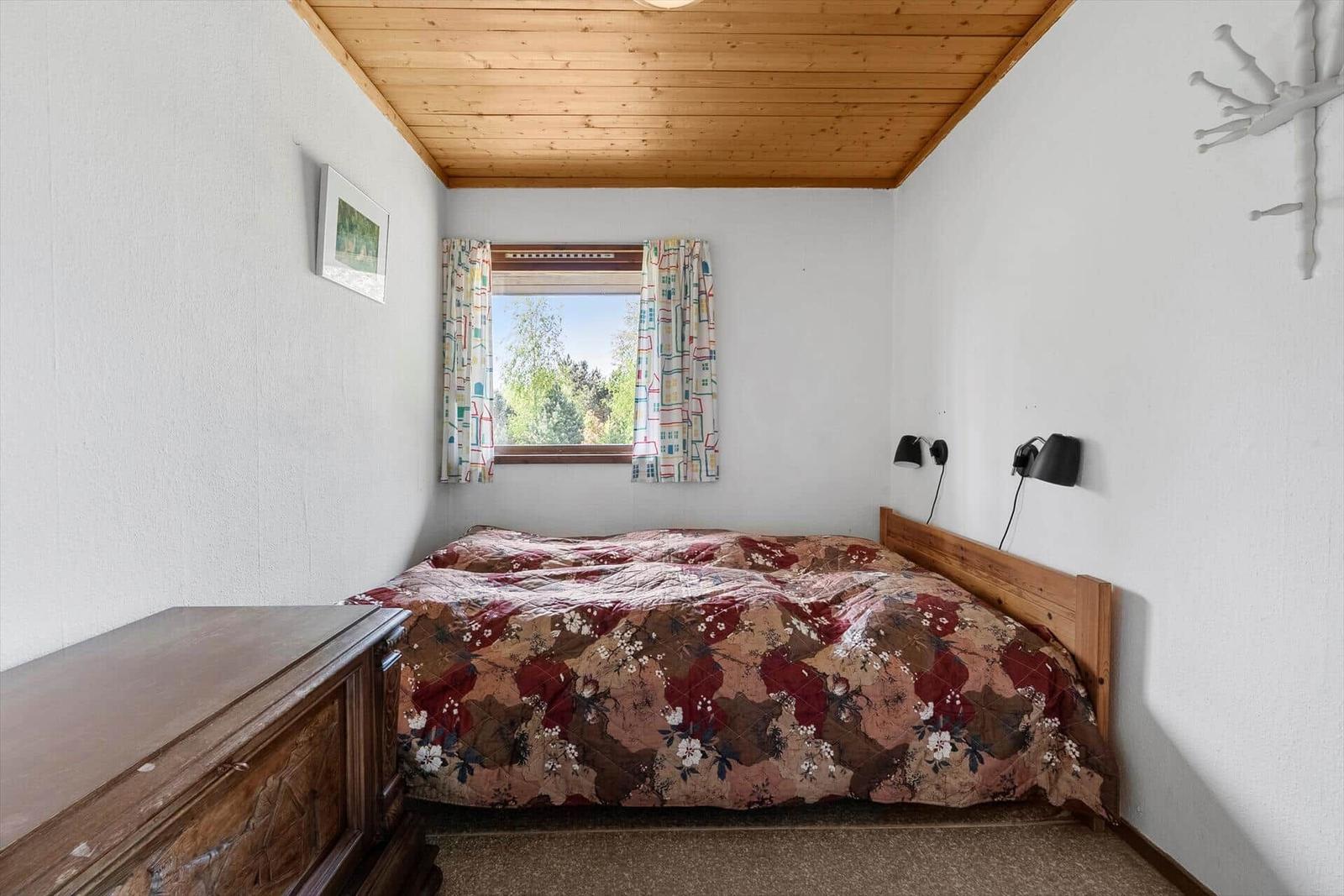 Schlafzimmer mit Holzdecke, Bett mit gemusterter Bettdecke und Fenster mit Vorhängen.