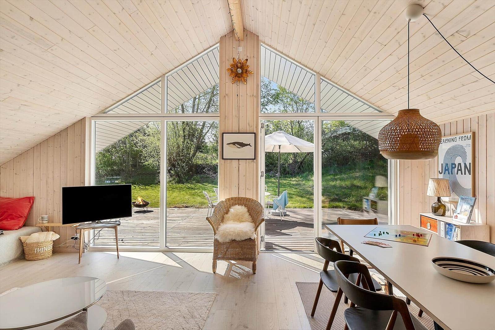 Wohnraum mit Holzwand, großem Fenster und Essbereich. Draußen Terrasse mit Sonnenschirm.