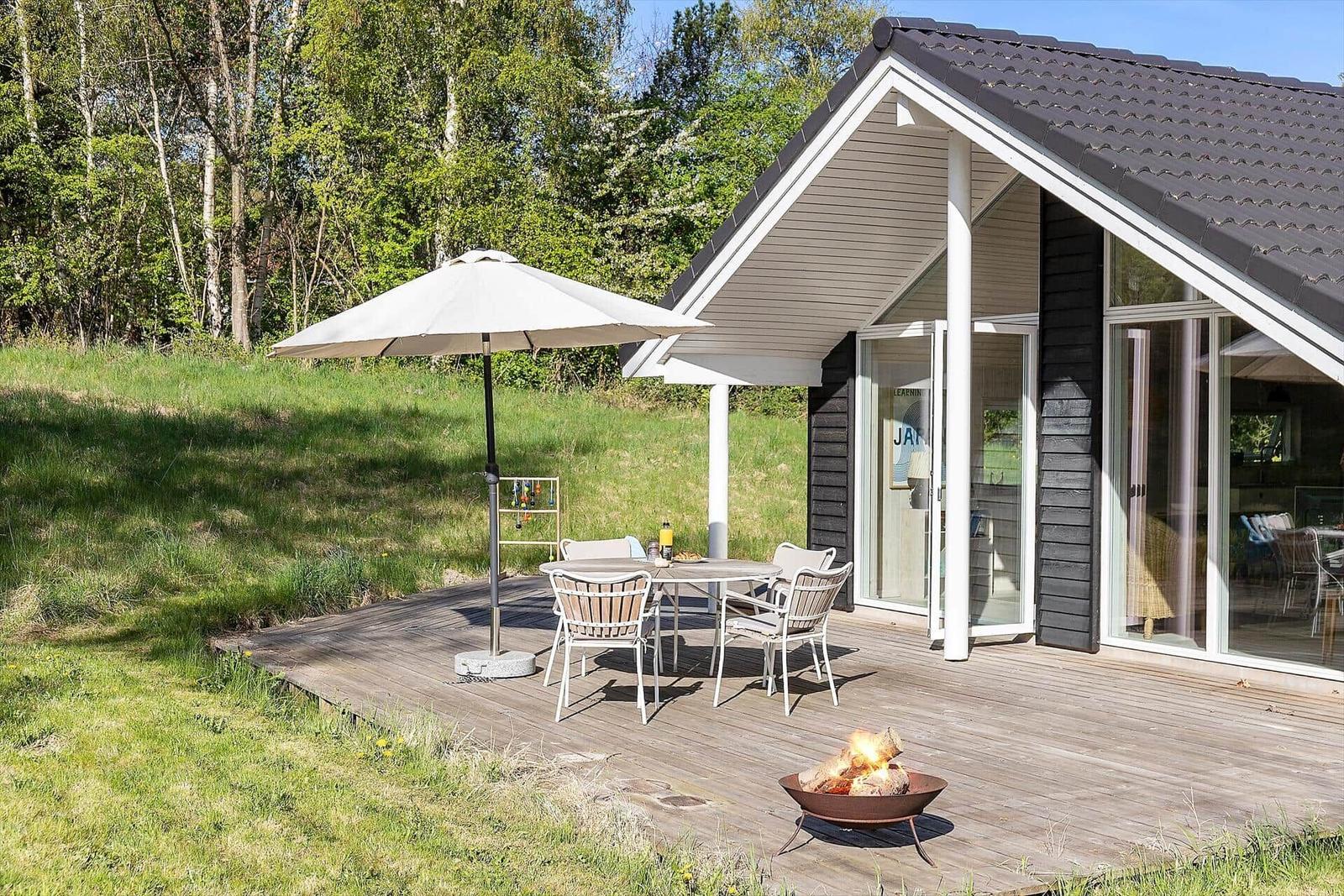 Terrasse mit Tisch, Stühlen, Sonnenschirm und Feuerkorb vor einem Haus mit Glasfront.