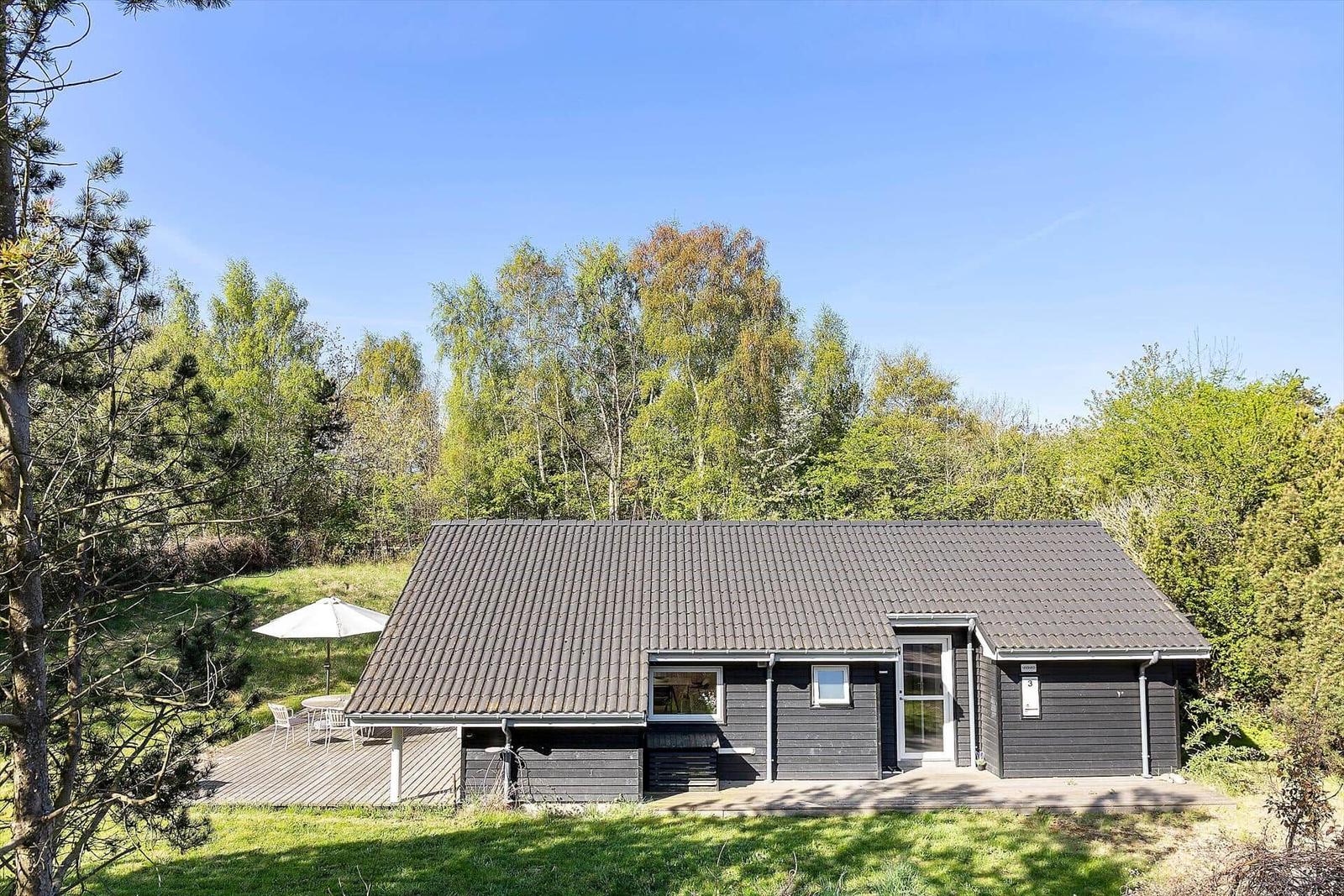 Ein dunkel verputztes Haus mit Holzveranda und Garten. Umgeben von Bäumen und einem weißen Sonnenschirm.