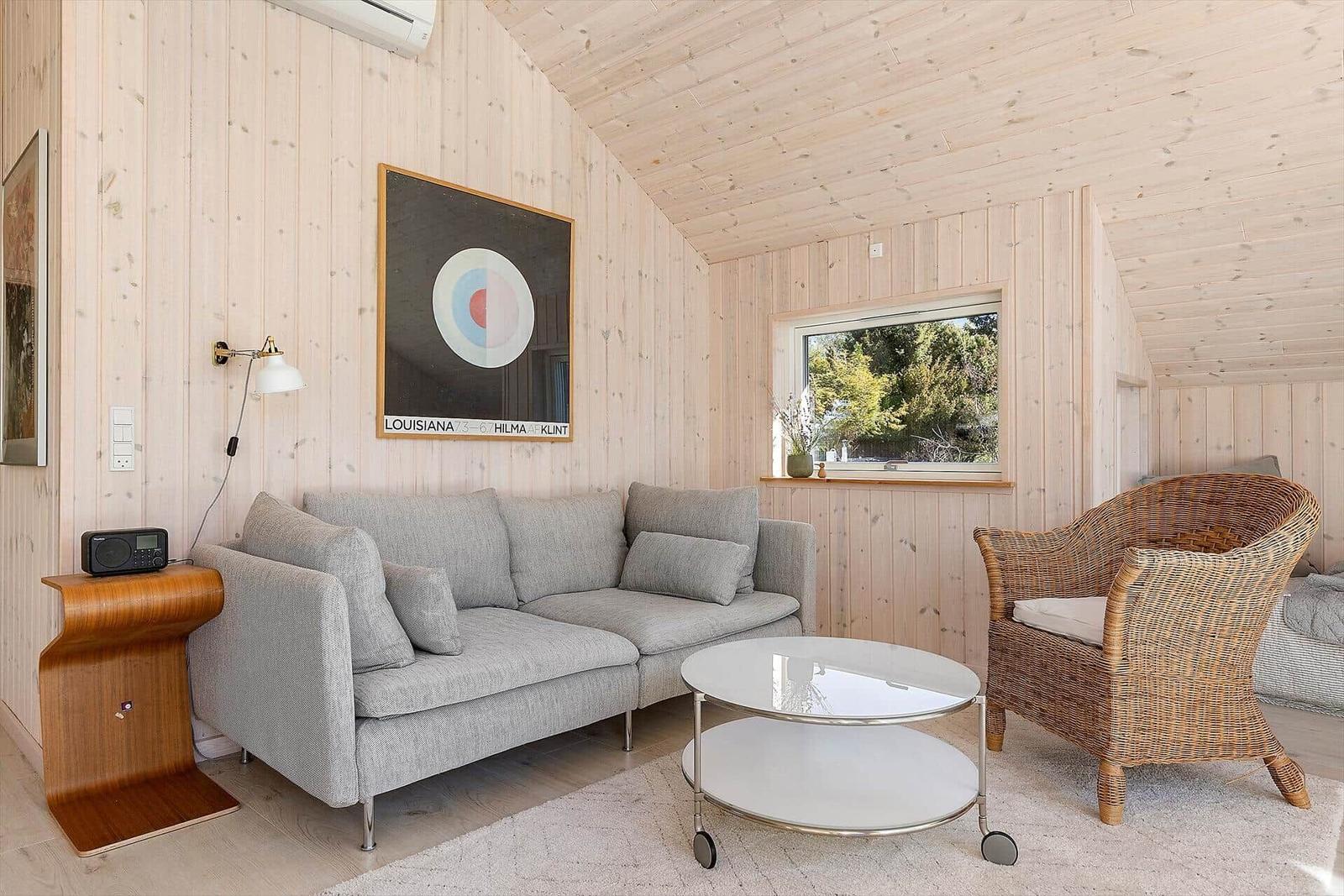 Wohnzimmer mit Sofa, Rattan-Stuhl und Glas-Tisch. Holzwände und Fenster mit Blick auf Grün.