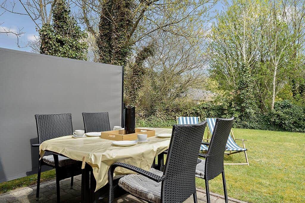 Gartenbereich mit Tisch, Stühlen und Sonnenschutz. Hintergrund: Bäume und Rasen.