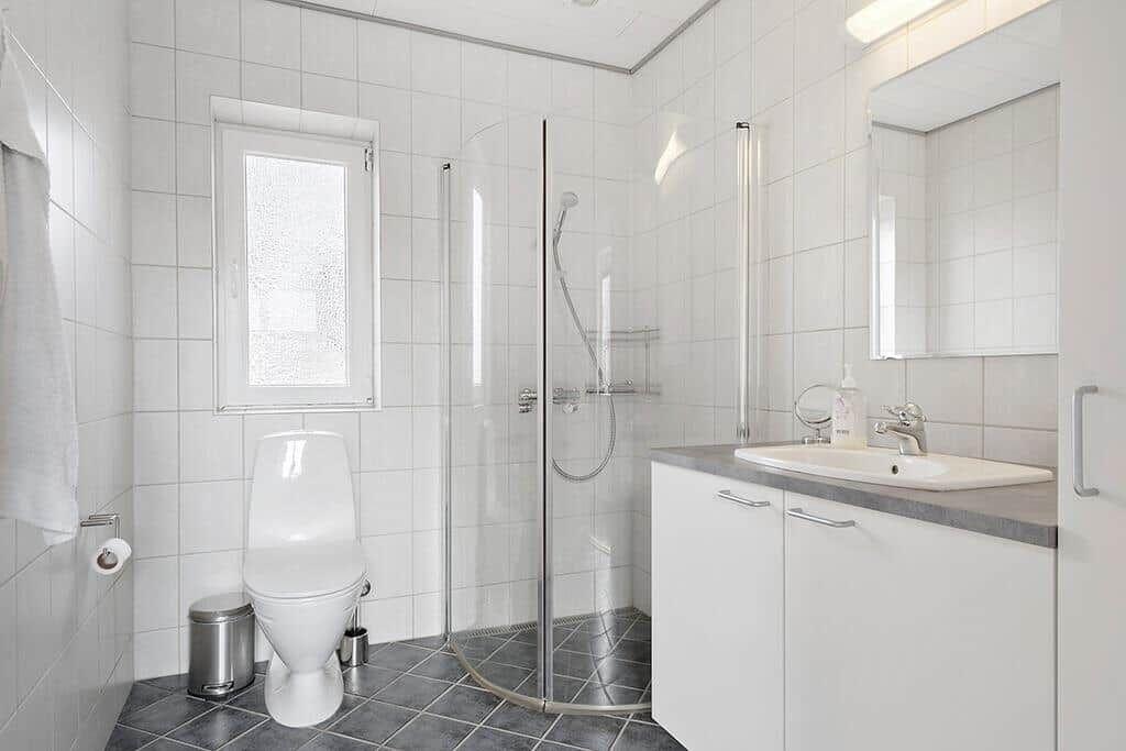 Badezimmer mit Dusche, Waschbecken und Toilette. Weiße Fliesen und graue Bodenfliesen.