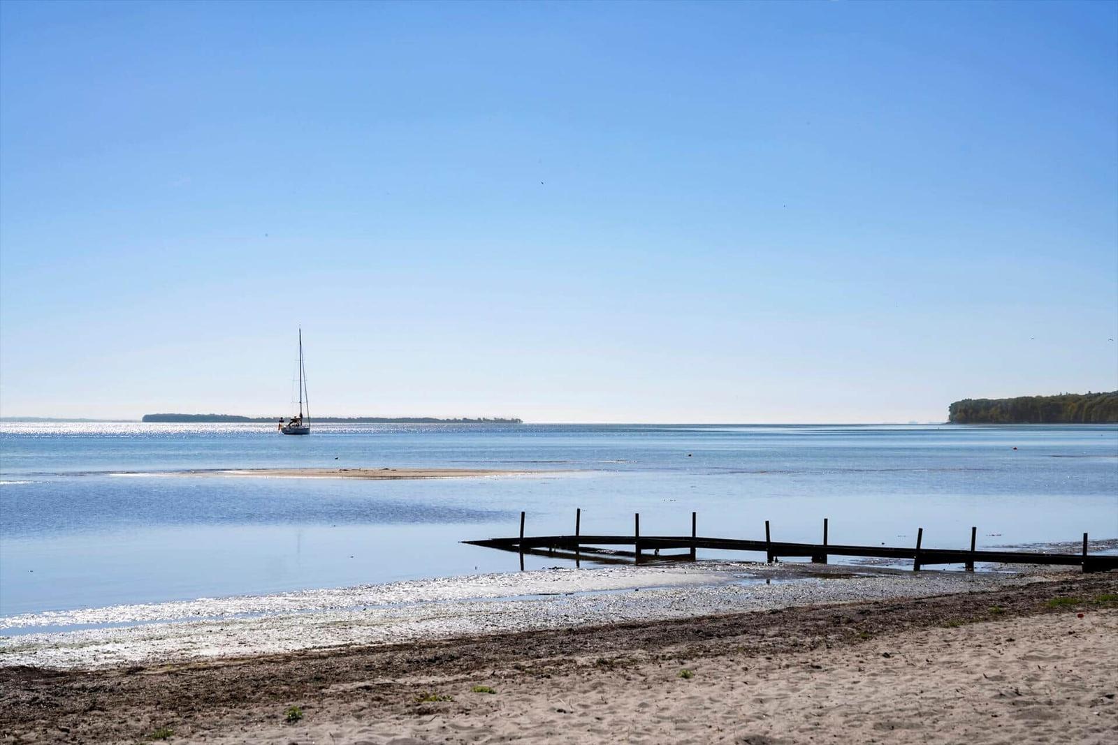 A sailboat rests on calm water, with a sandy beach and wooden pier visible.