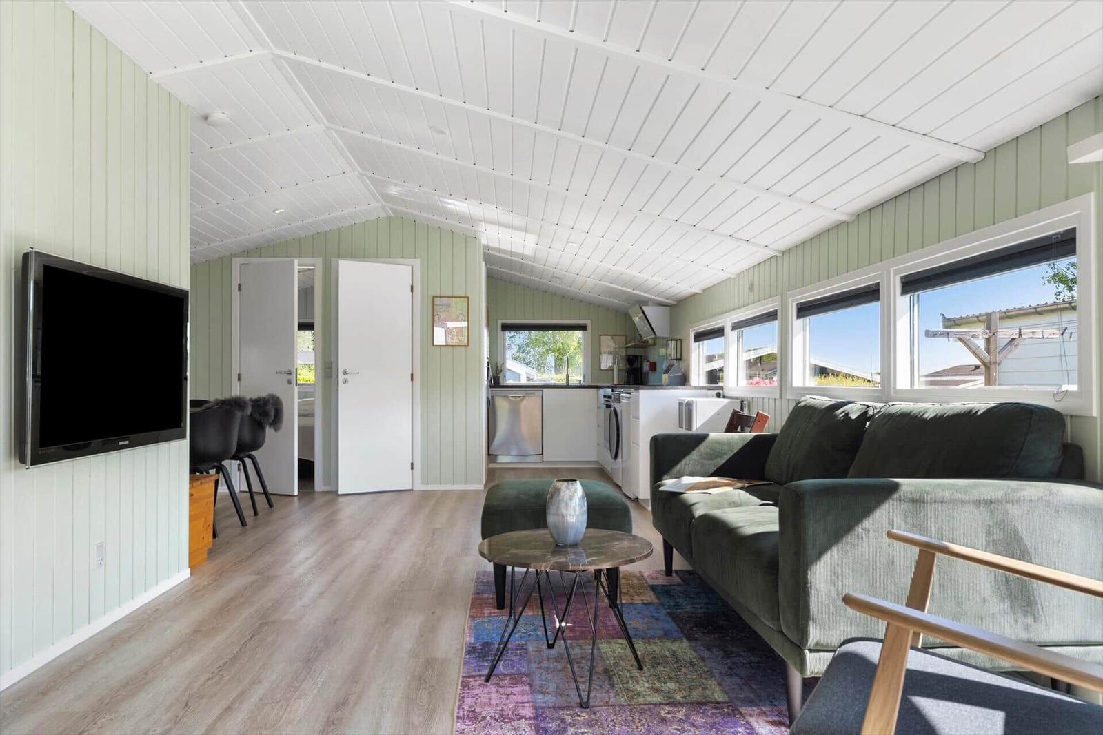 Living area with sofa, TV, and kitchen area. Wooden floor and green walls.