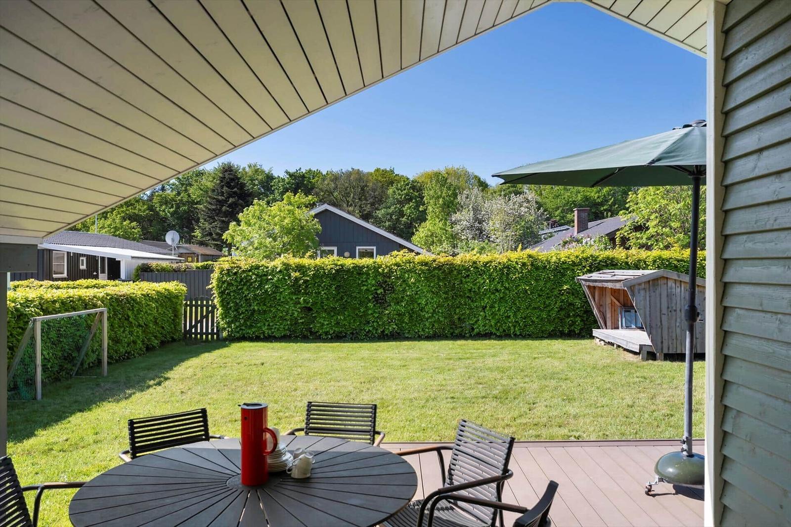 Terrace with table, chairs, and umbrella. Lawn and hedge in the background.