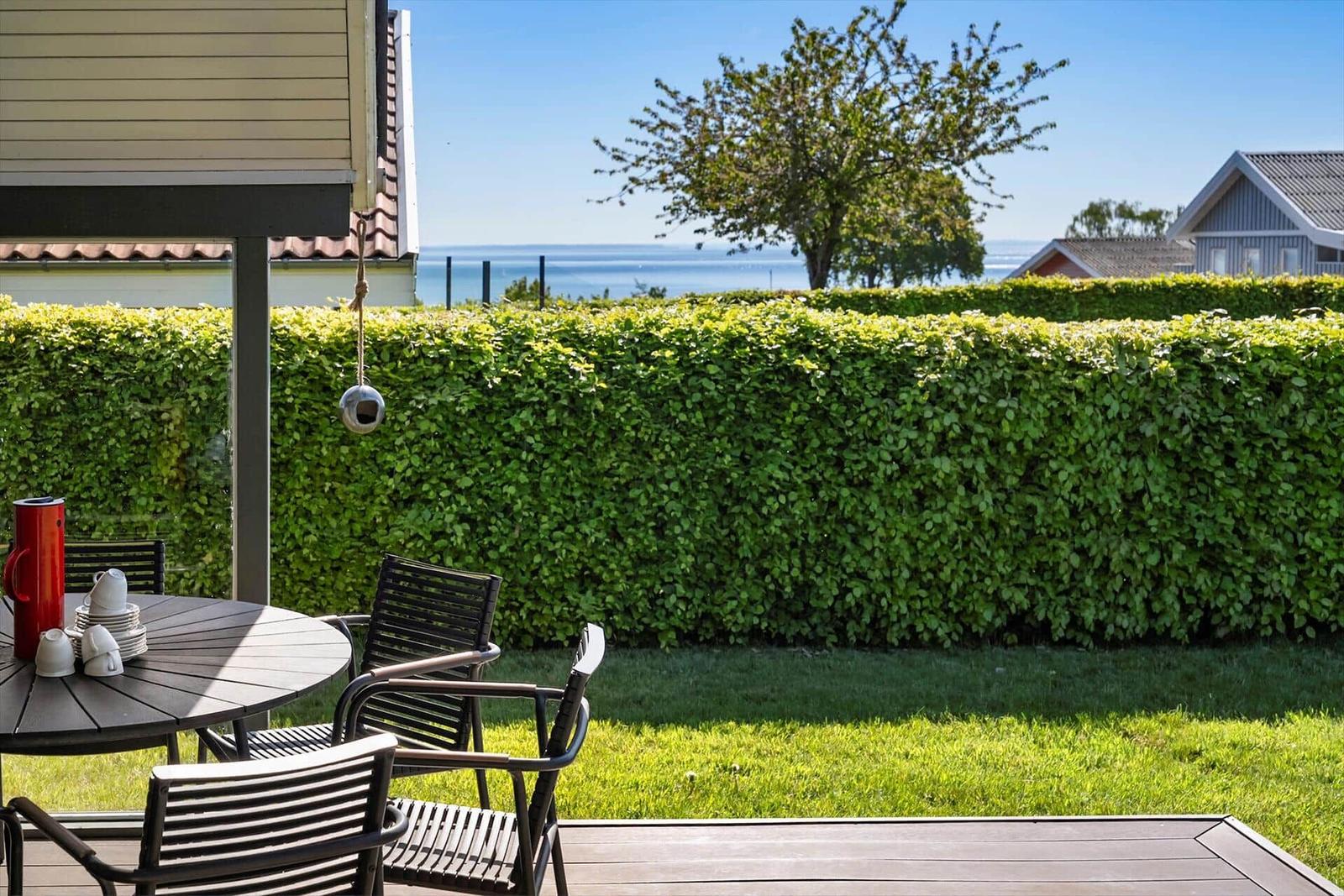 Deck with table and chairs, view of green hedge and sea.