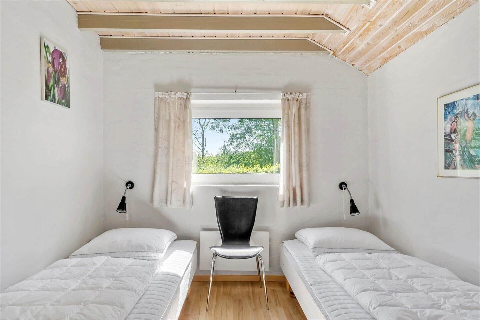Two single beds, window with curtains, black chair, wooden ceiling, wall art.