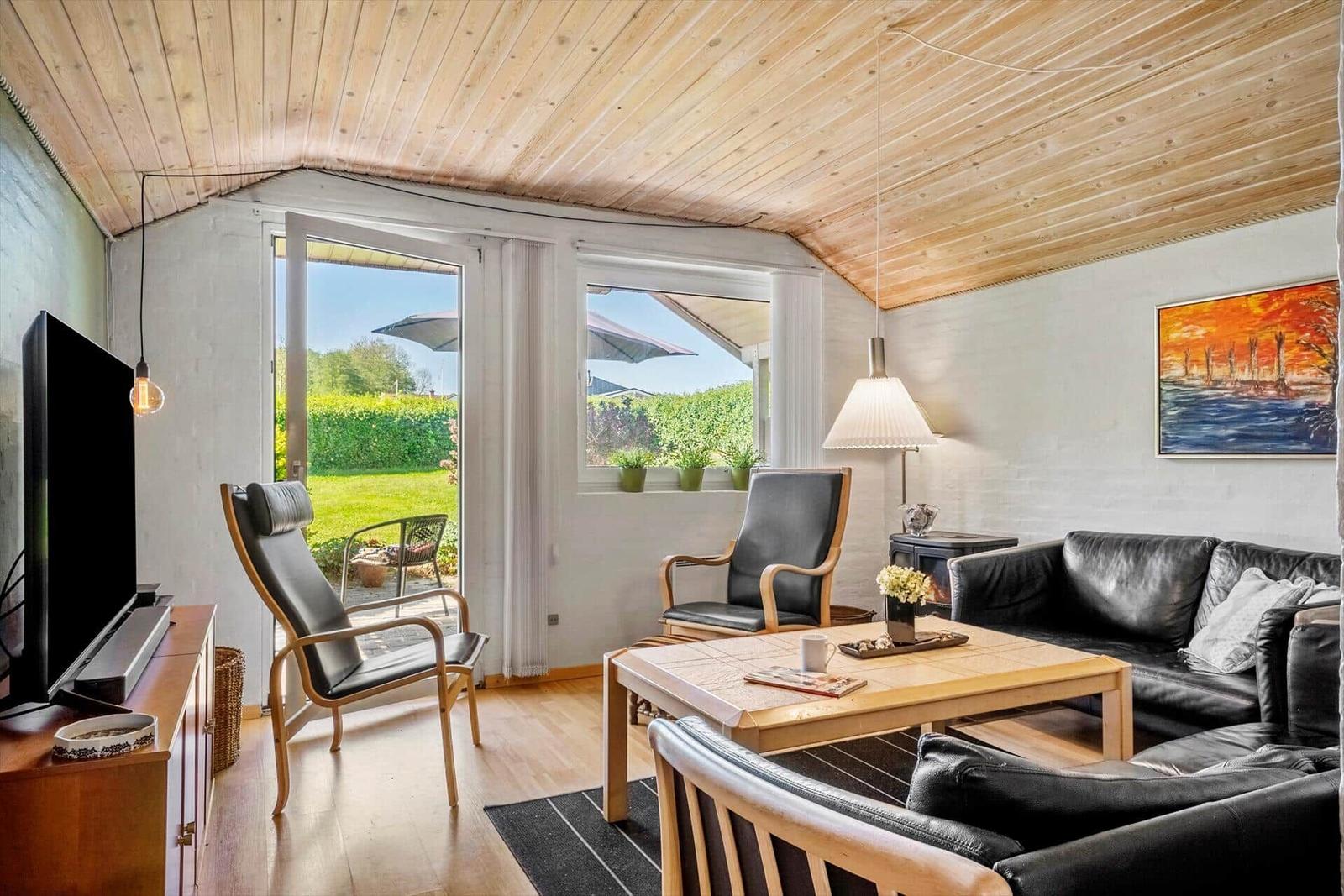Living room with wooden ceiling, sofa, chairs, and TV. View of garden through open door.