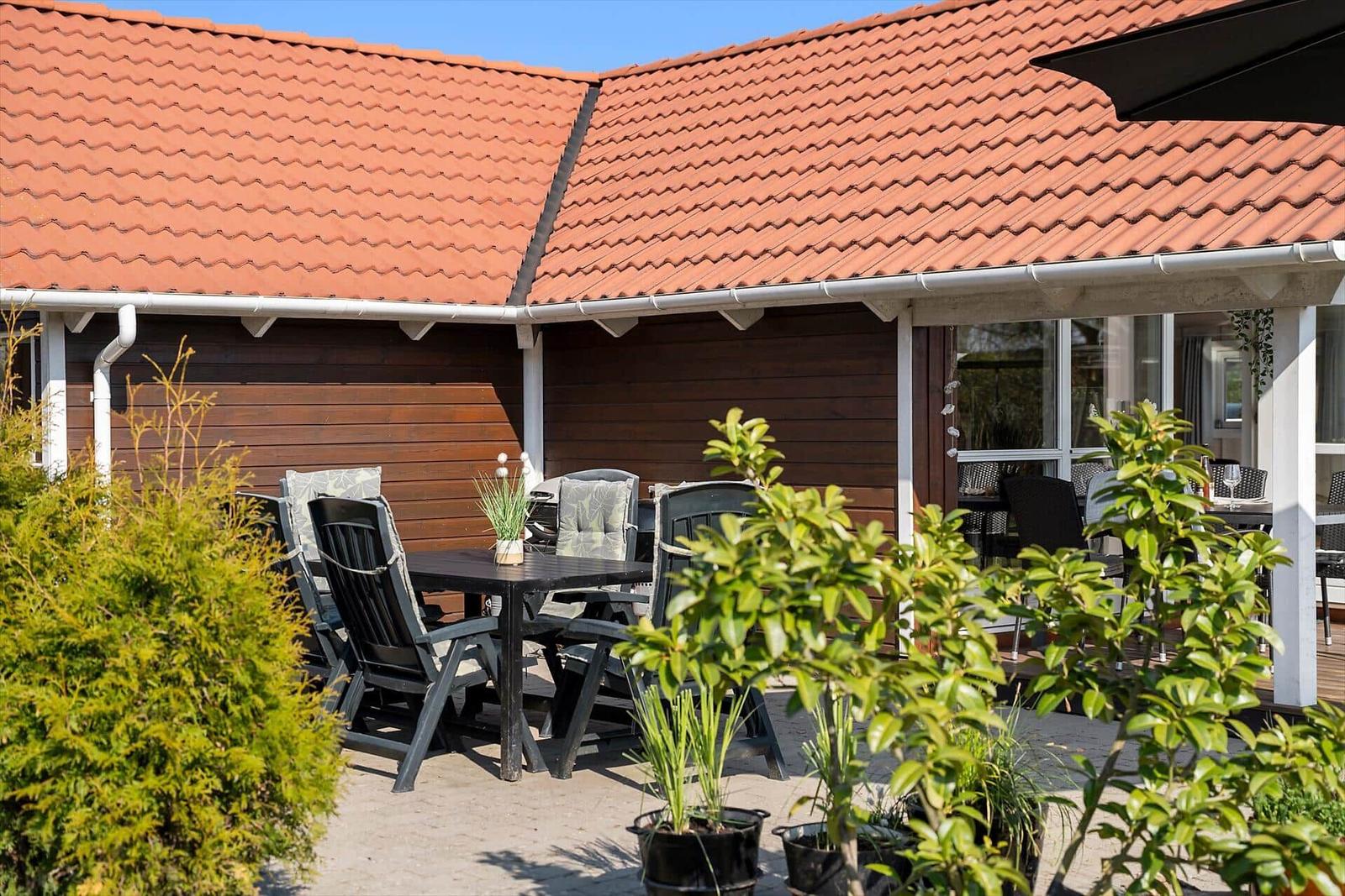Terrace with table, chairs, and plants in front of a house with red roof.