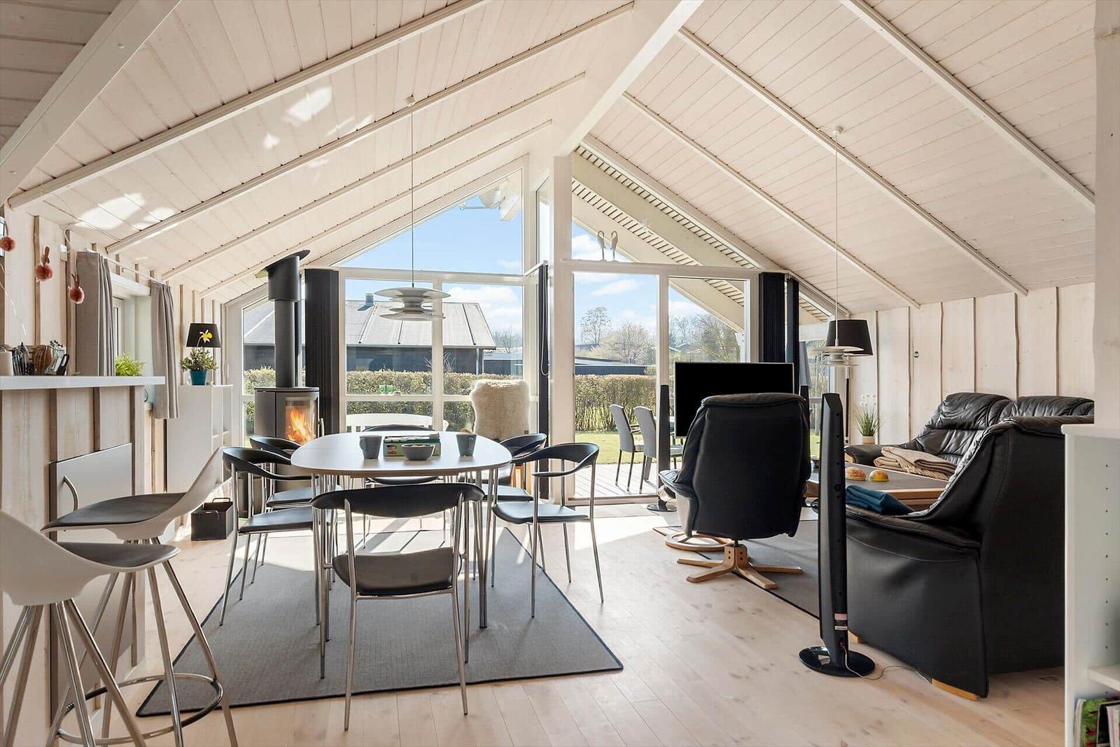 Living area with dining table, sofa, and fireplace. Large windows overlook the outdoor area.