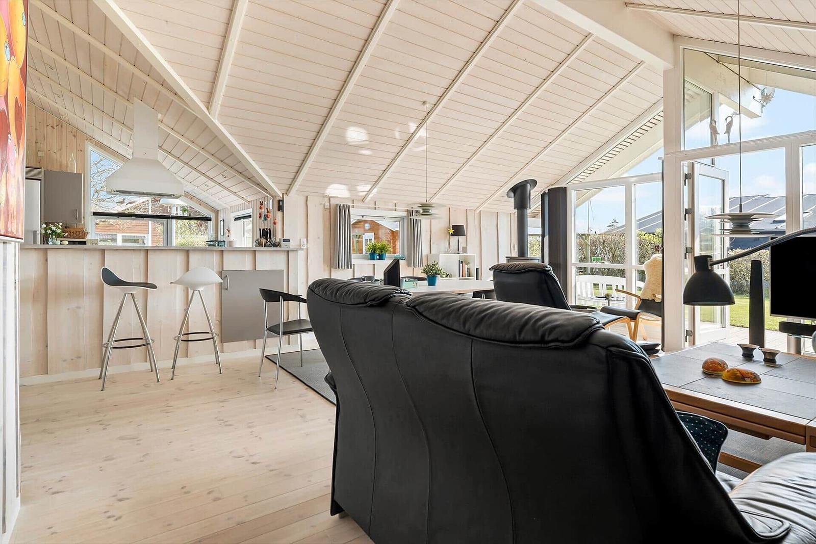 Living area with kitchen, sofa, and table. Large windows and wooden ceiling.