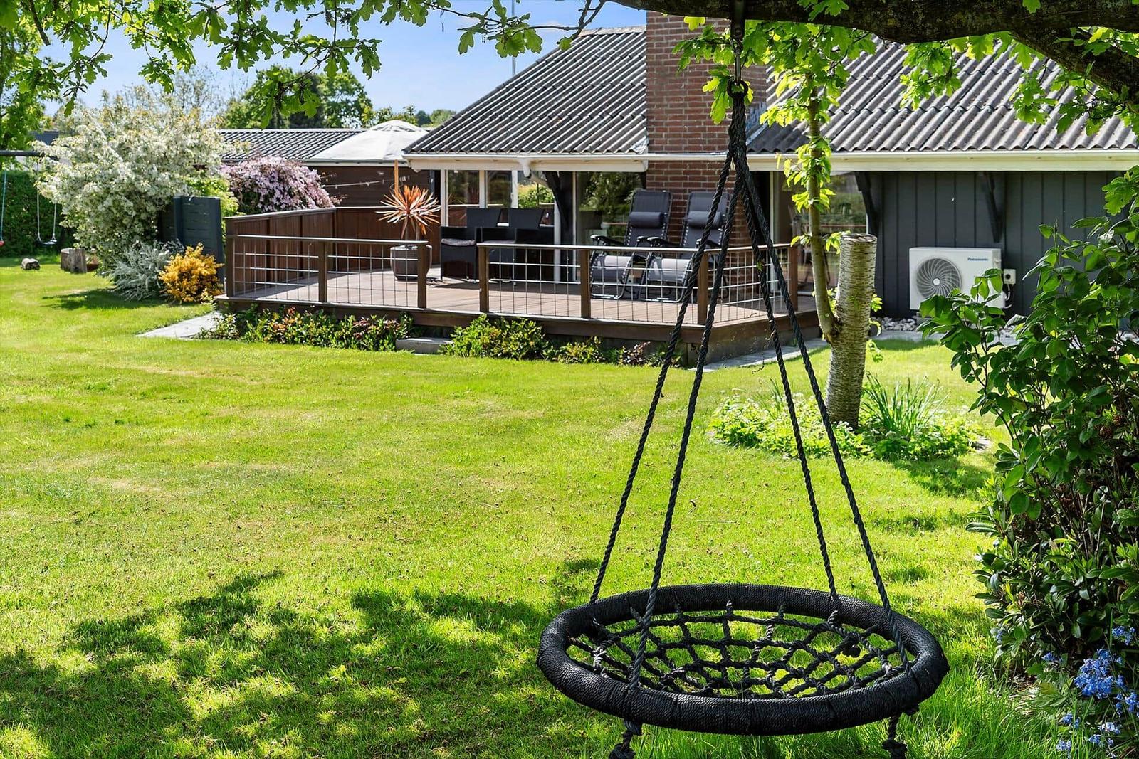 Schwarzes Seil-Schaukelbett hängt unter einem Baum im Garten. Im Hintergrund eine Terrasse mit Sitzgelegenheiten.