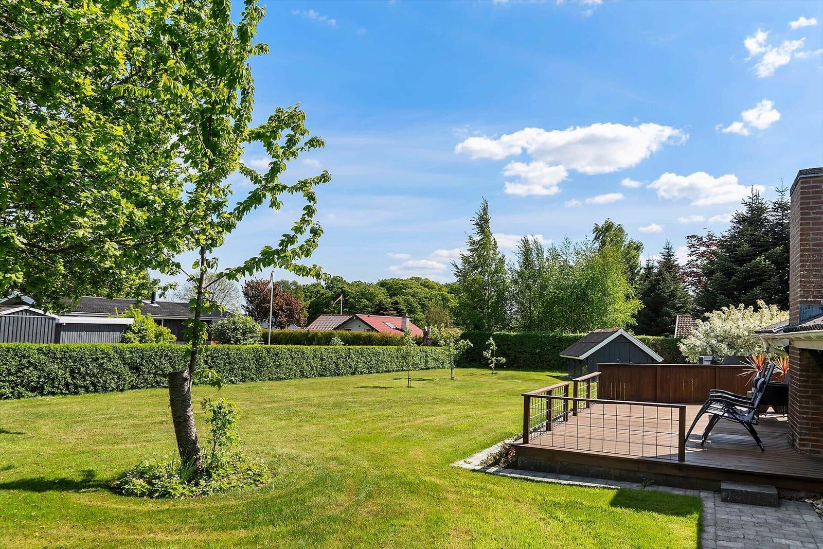 Hinterhof mit Terrasse, Rasen und Bäumen unter blauem Himmel.
