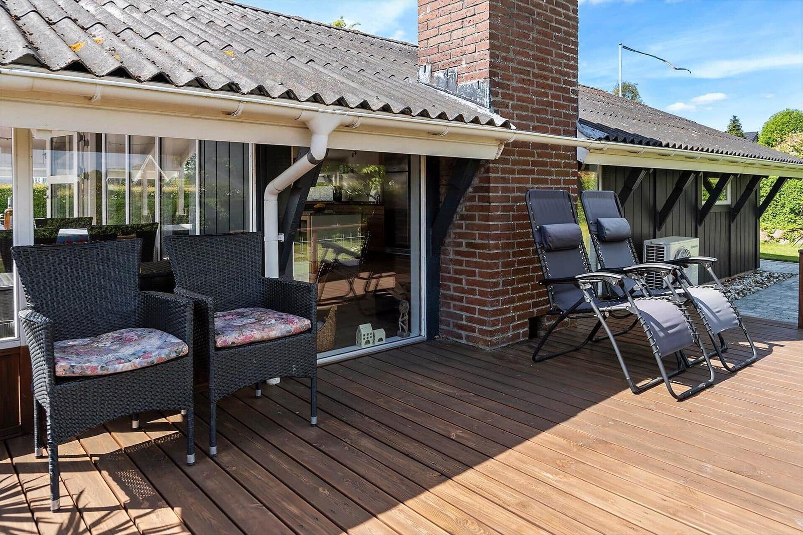 Terrasse mit zwei Sesseln und zwei Liegestühlen. Holzdielen und Glasfront sichtbar.