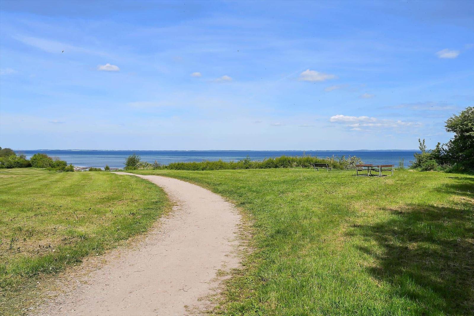 Eine Straße führt durch eine grüne Wiese zum Meer. Es gibt Bänke und einen Tisch.