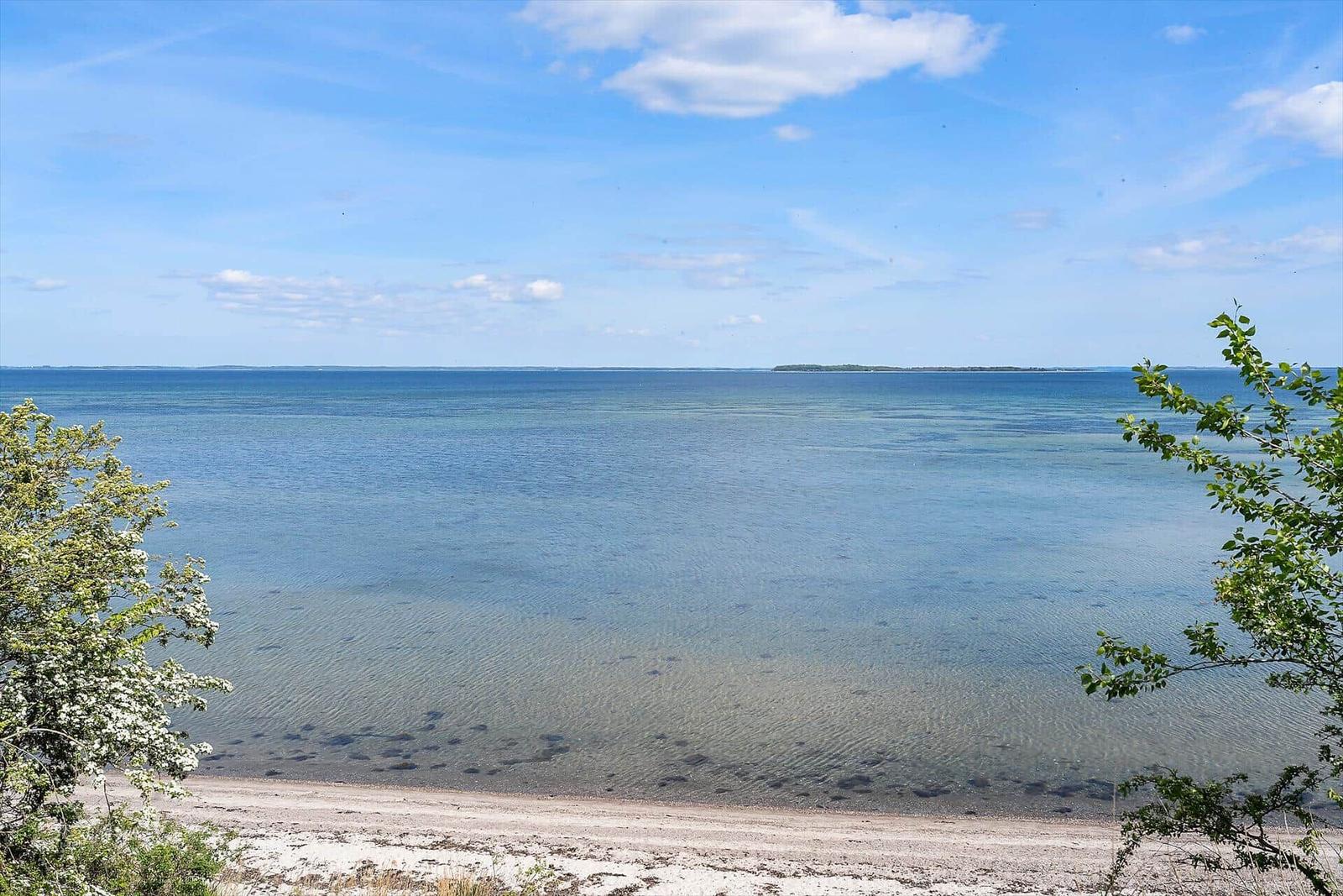 Strand mit klarem Wasser und blauem Himmel. Bäume im Vordergrund.