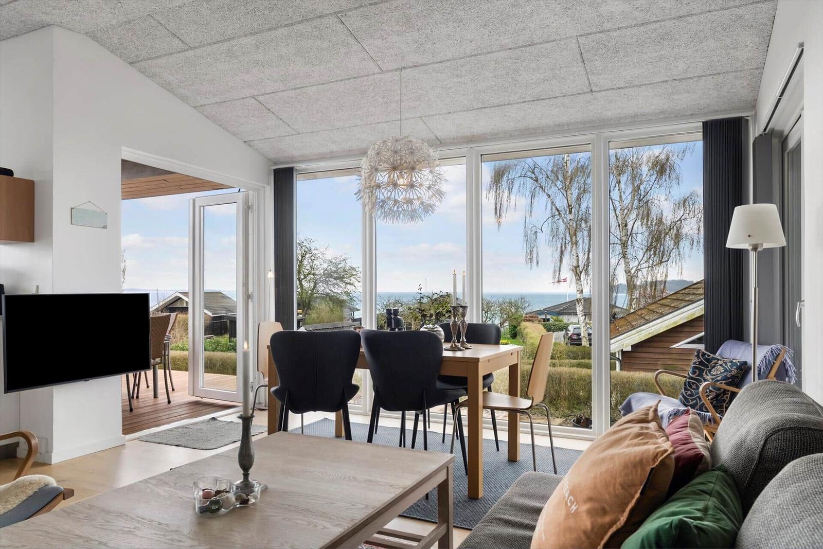 Living and dining area with view of the lake. Large windows, table, sofa, and TV.