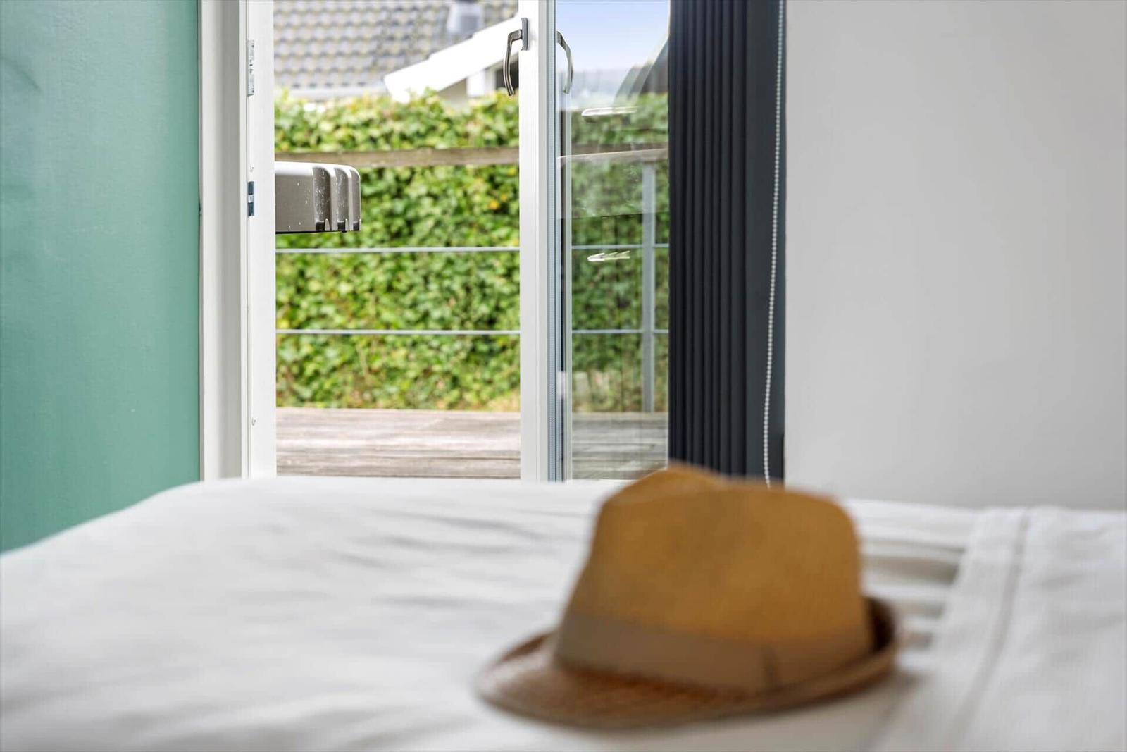 A bed with a hat in the foreground. Through the window, a terrace and greenery are visible.