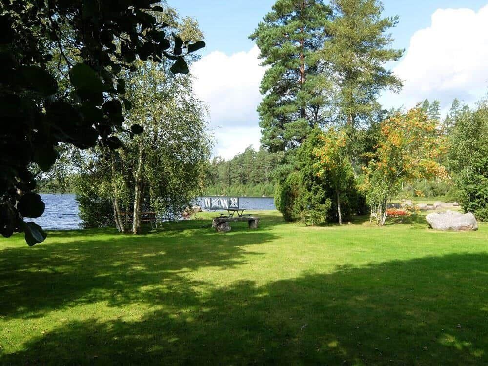 Grüner Rasenbereich mit Bäumen und Blick auf See. Picknicktisch und Stein im Vordergrund.