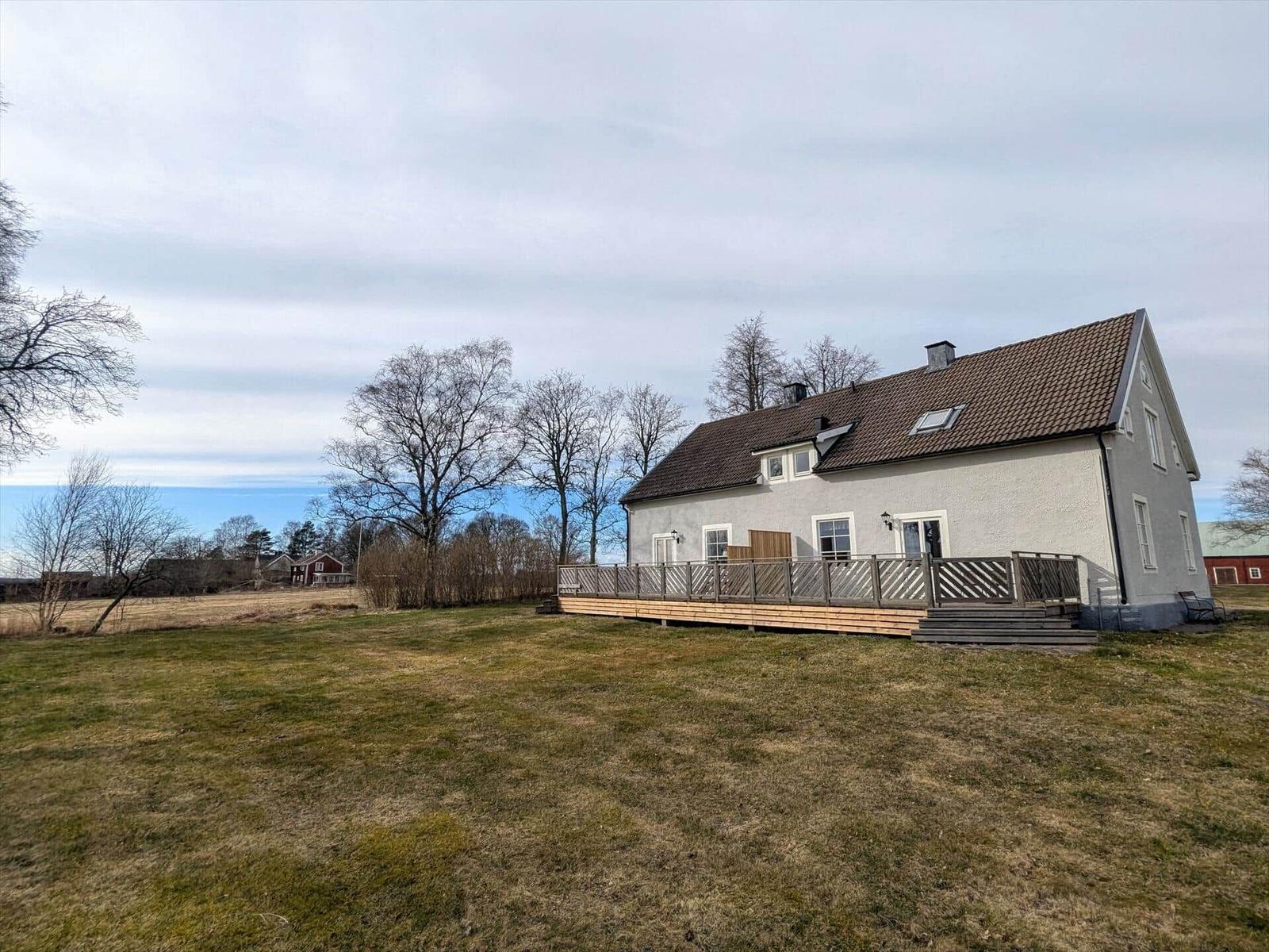 Ein zweistöckiges Haus mit Terrasse und Garten. Im Hintergrund sind Bäume und ein rotes Gebäude sichtbar.