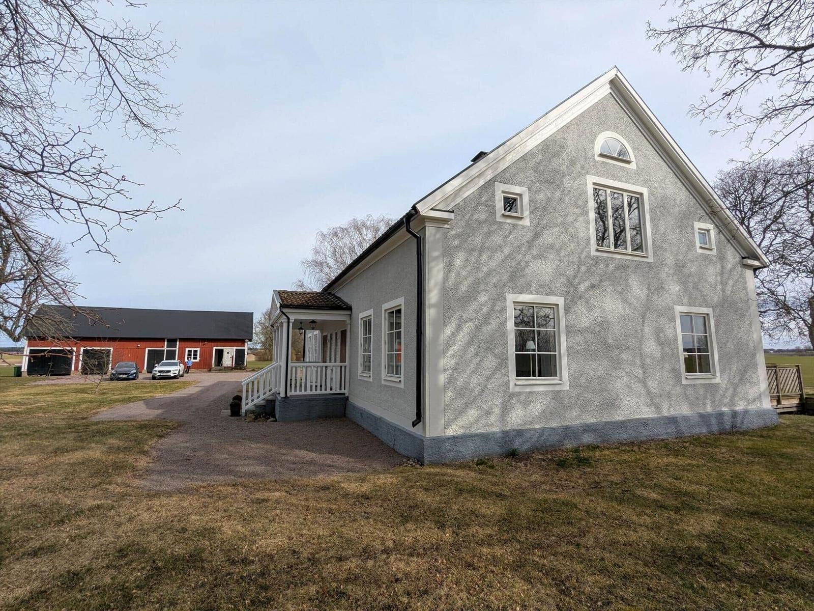 Ein graues Haus mit weißen Fensterrahmen und einer Veranda. Im Hintergrund steht ein roter Schuppen.