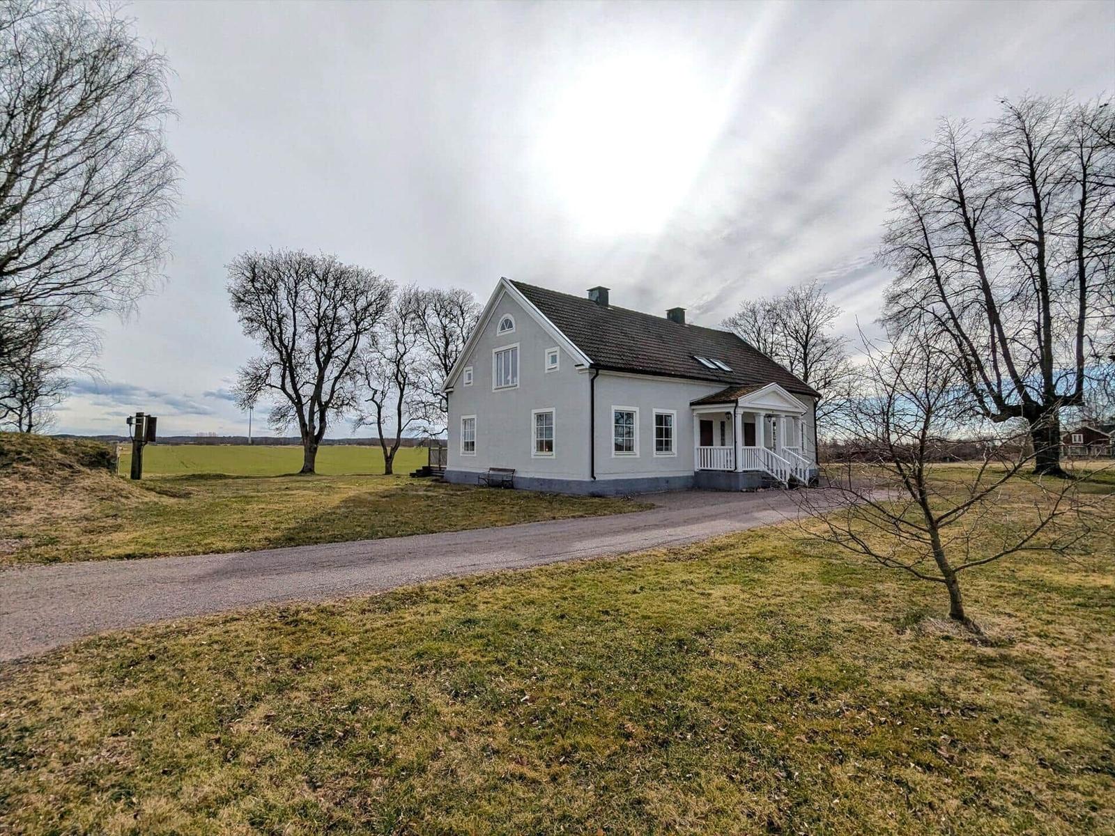 Ein zweistöckiges Haus mit weißer Fassade und Vordach. Umgeben von Gras und Bäumen.
