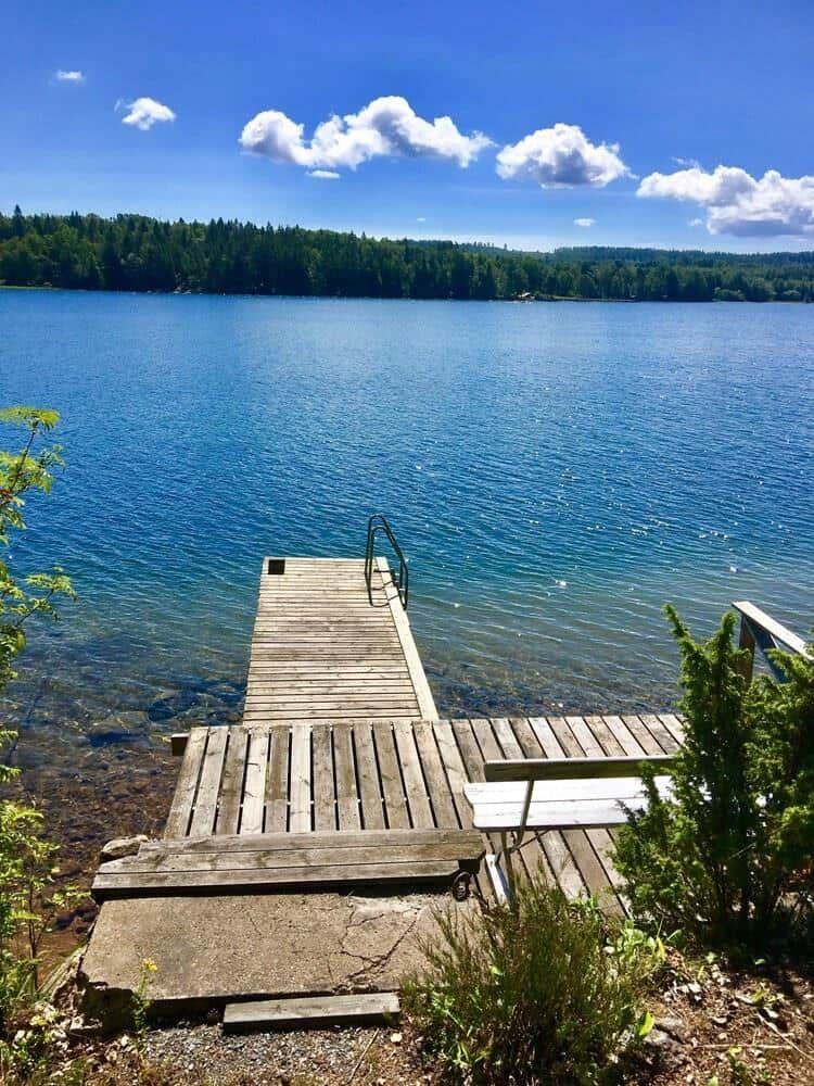 Holzsteg führt zu einem See mit klarer blauer Wasserfläche und grüner Waldlandschaft im Hintergrund.