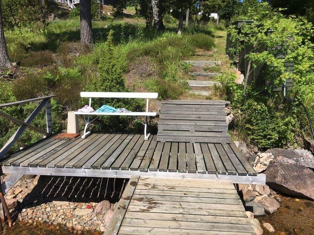 Holzbrücke mit Bank über Bach, umgeben von Grün und Steinen.