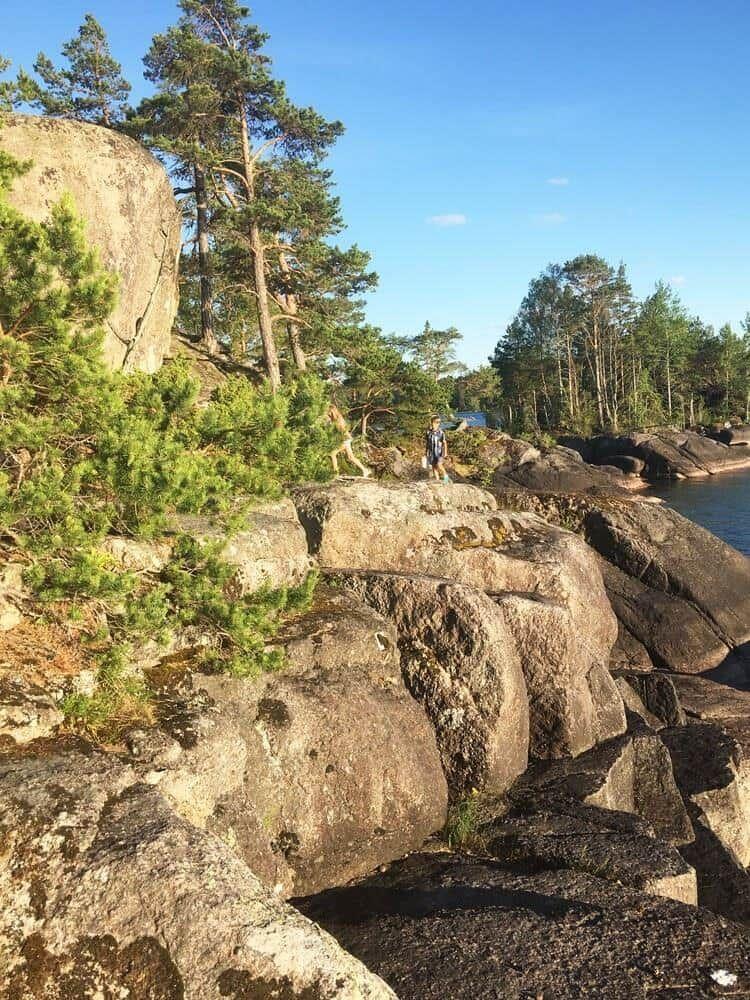 Person steht auf Felsen am Ufer mit Bäumen und klarem Himmel.