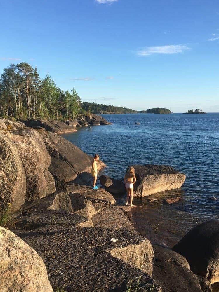 Zwei Kinder stehen auf Felsen am Wasser. Im Hintergrund sind Bäume und Inseln zu sehen.