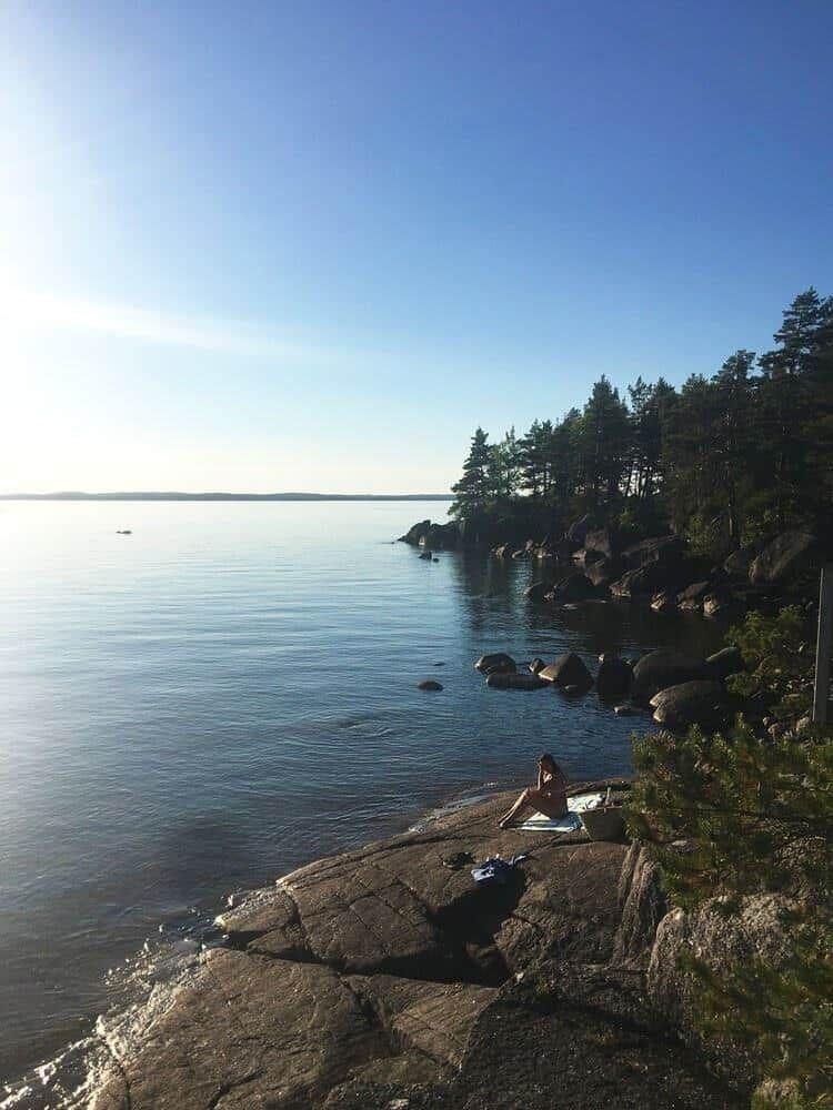 Person sitzt auf Felsen am ruhigen Wasser. Bäume und klarer Himmel im Hintergrund.