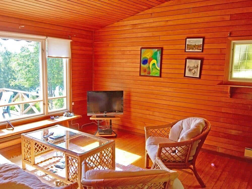 Wohnzimmer mit Holzwand, Fernseher, Sessel und Glas-Tisch. Fenster mit Blick auf Balkon.
