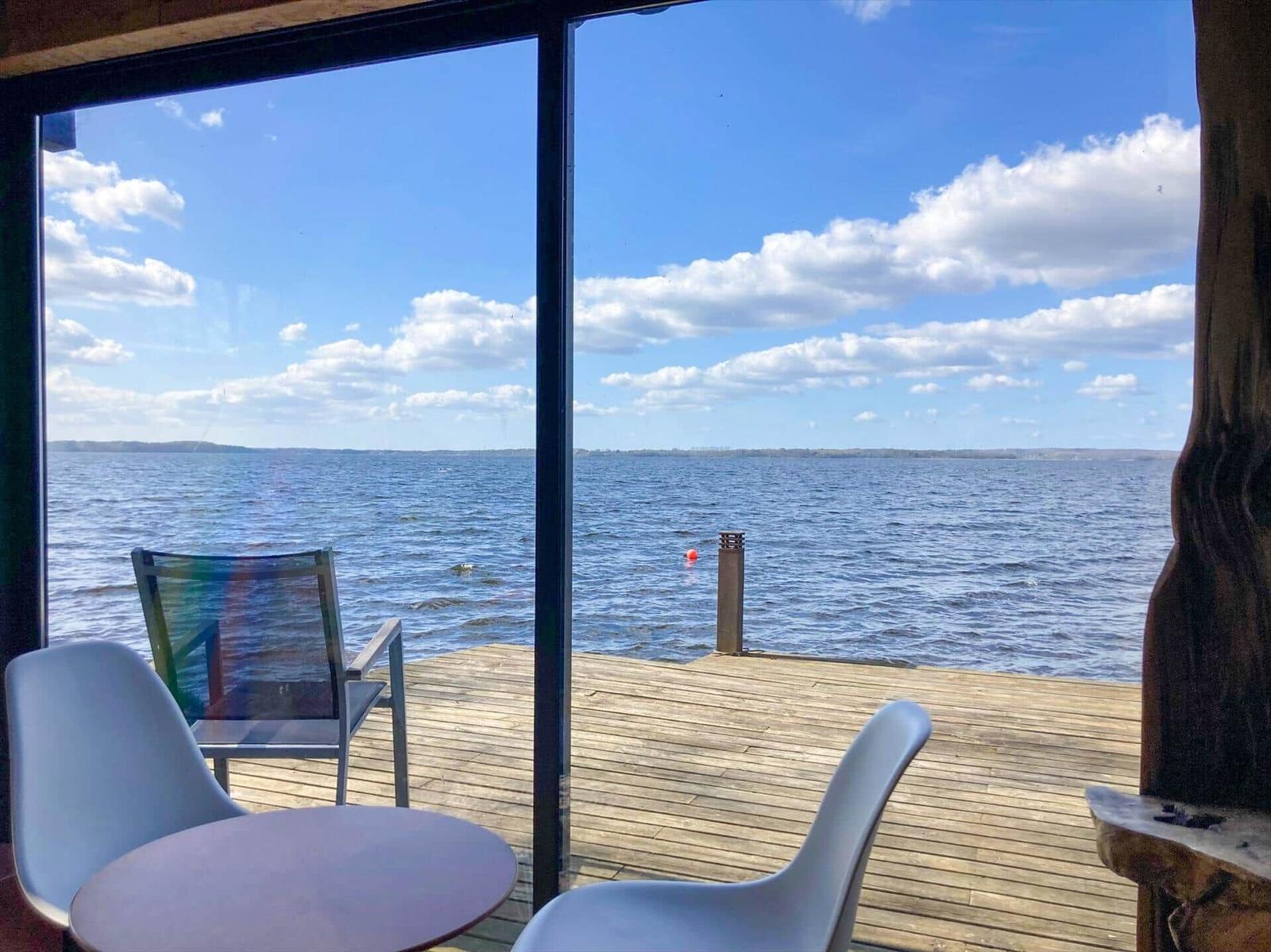 Von innen betrachtet, ein Blick auf einen Holzsteg mit Stühlen und Tisch am Wasser.