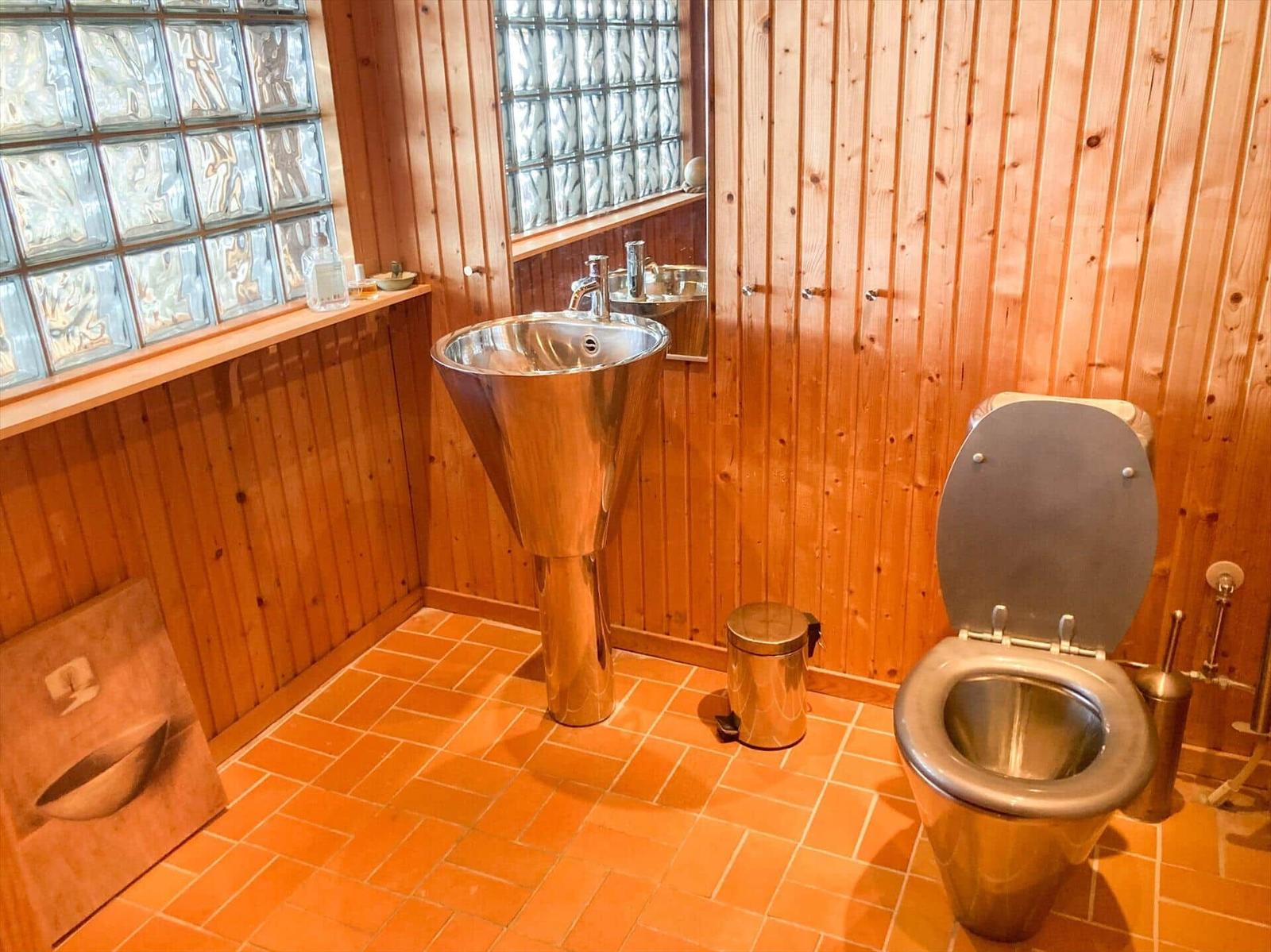 Bad mit Holzwand, Glasblockfenster, Edelstahlwaschbecken und Toilette auf Terrakottboden.