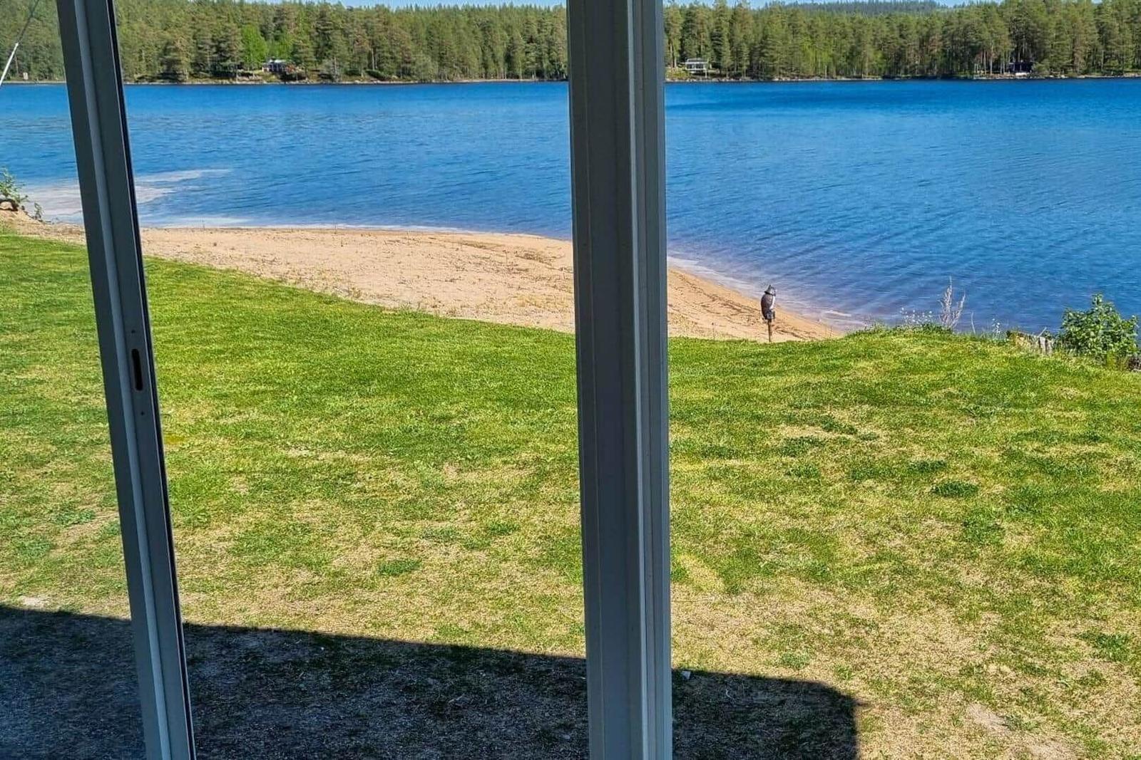 Blick auf einen See mit Sandstrand und Wald im Hintergrund durch eine Glasschiebetür.