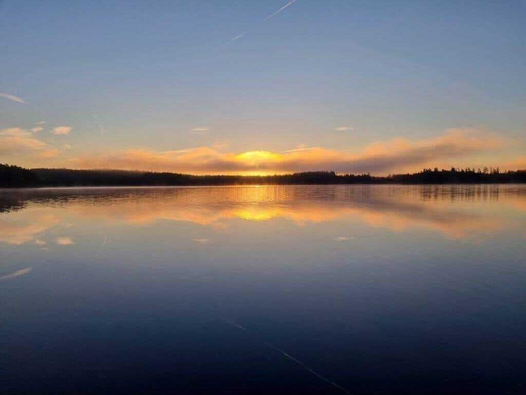 Sonnenuntergang über einem ruhigen See mit Spiegelung im Wasser und Wald am Horizont.
