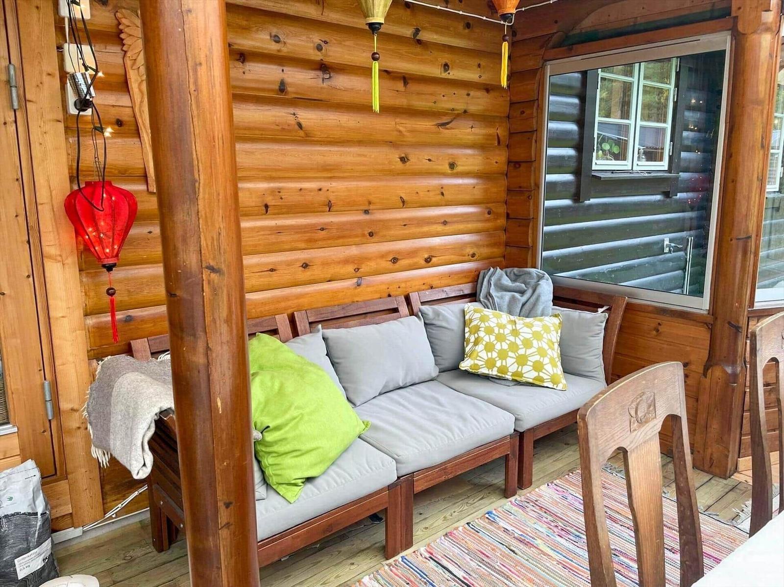 Holzveranda mit Sitzbank, Kissen und Teppich. An der Wand hängt eine rote Laterne.