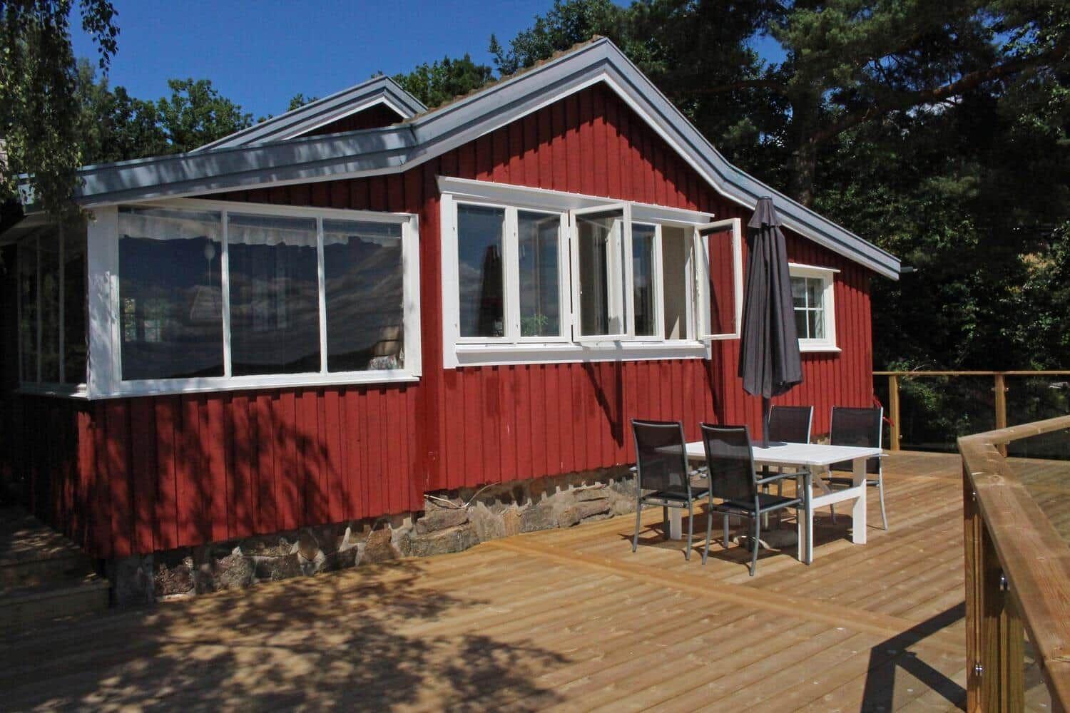 Rotes Holzhaus mit Terrasse und Esstisch. Umgeben von Bäumen unter blauem Himmel.