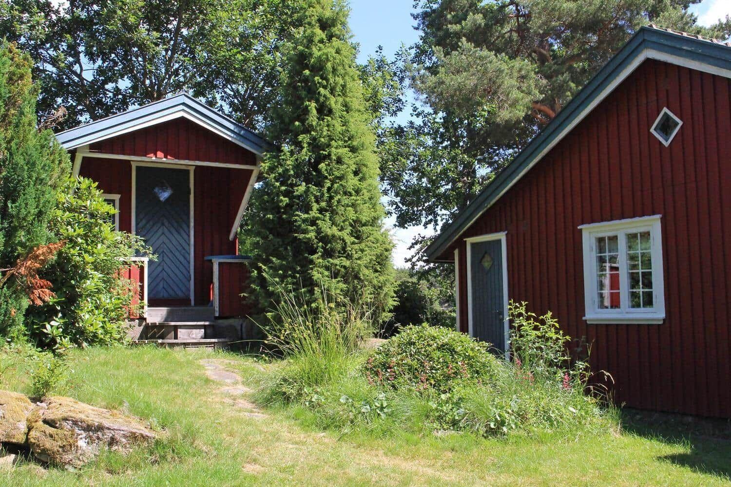 Zwei rote Holzhäuser mit weißen Fensterrahmen stehen im Gras zwischen Bäumen.