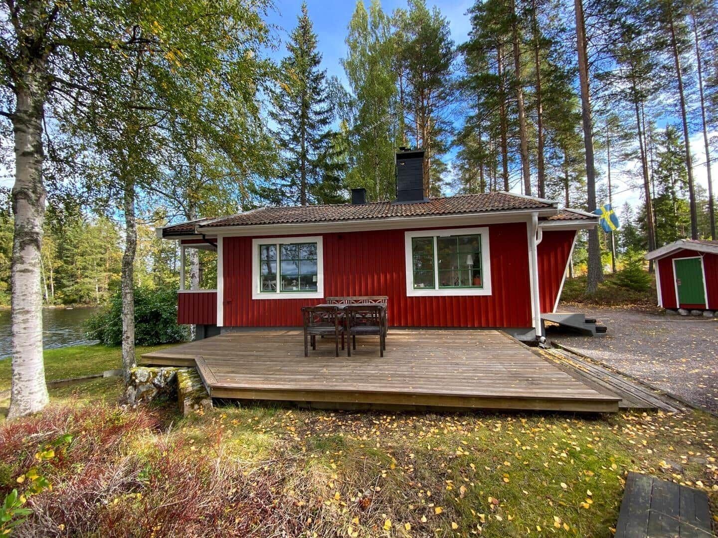 Rotes Holzhaus mit Terrasse am See, umgeben von Bäumen und einem kleineren Anbau.