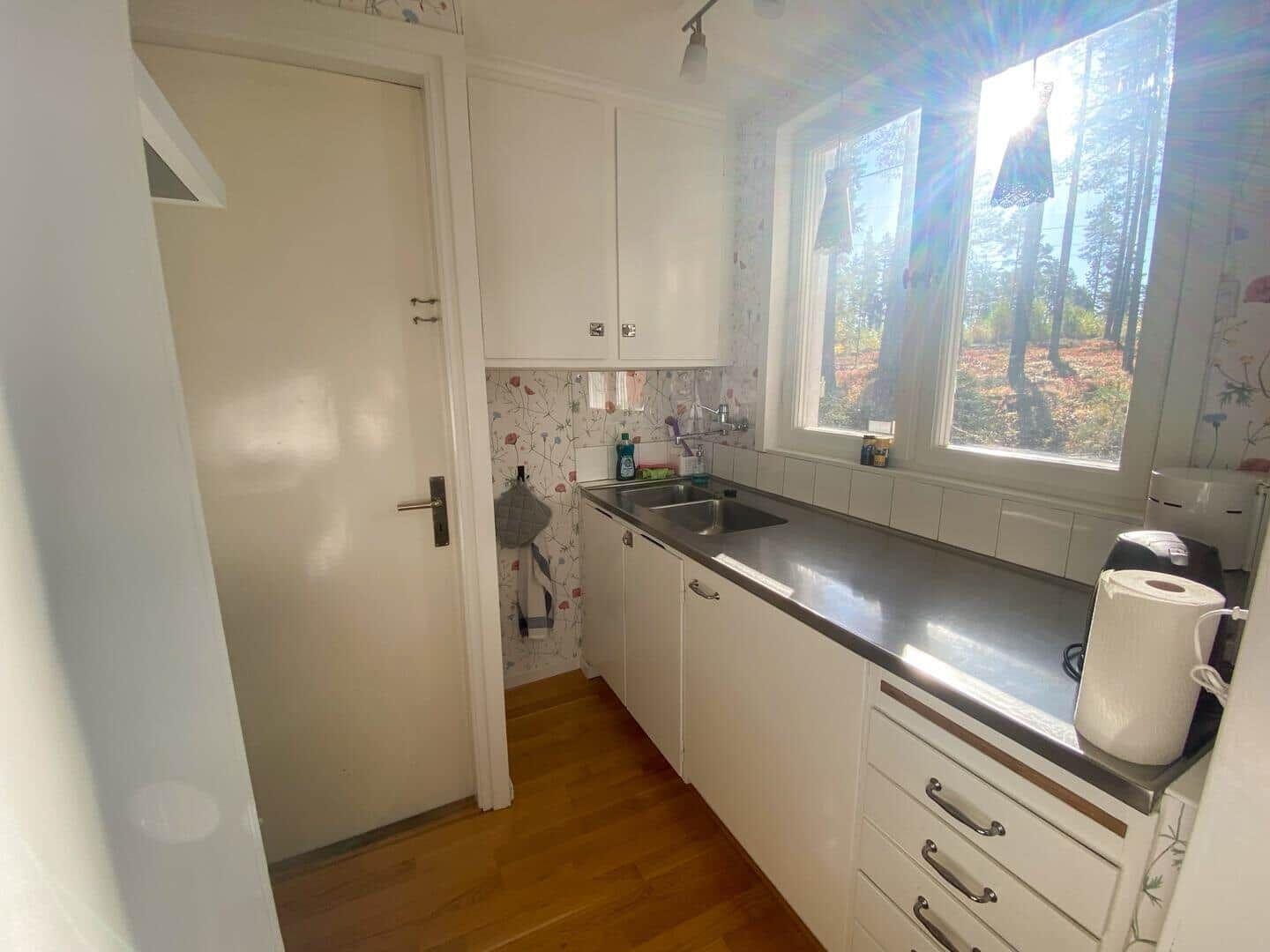 Küche mit Fensterblick auf Wald, weiße Schränke und Edelstahlarbeitsplatte.