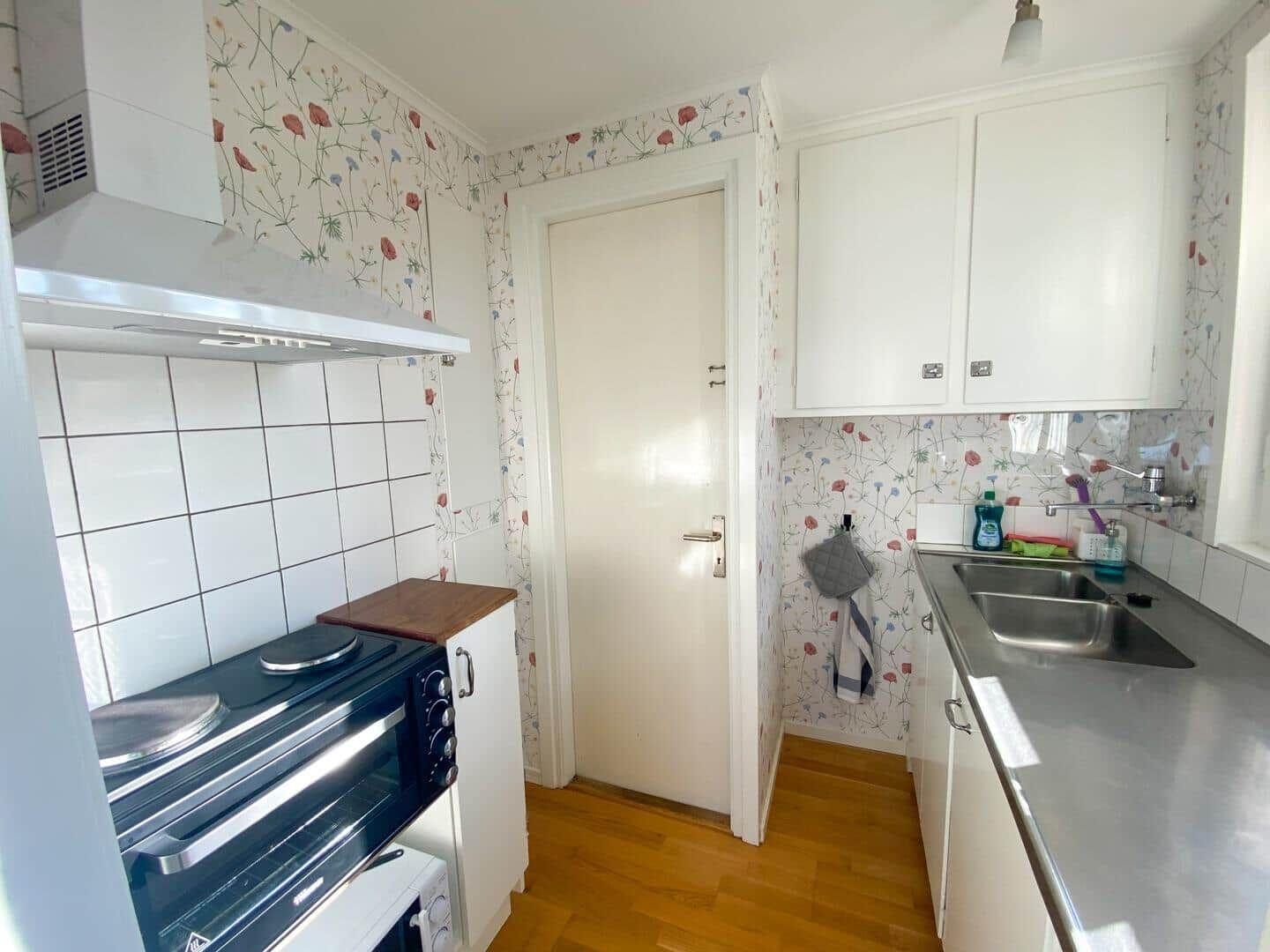 Küche mit weißen Schränken, Spüle und Herd. Wand mit Blumenmotiv und Holzboden.