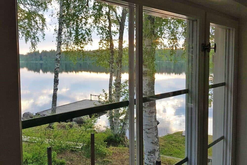 Blick auf ruhigen See mit Bootssteg und Birken durch große Fenster.
